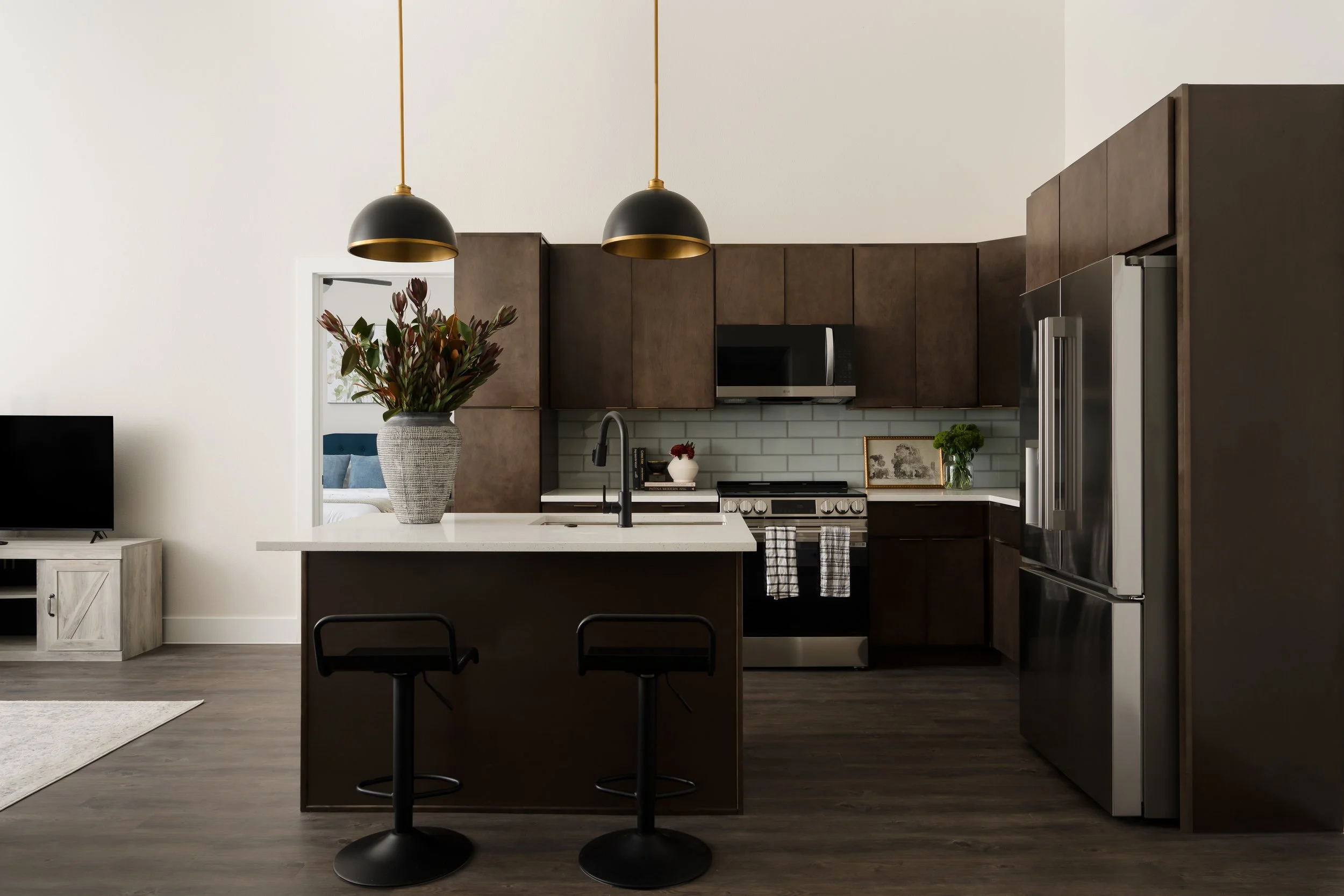 Modern apartment kitchen at The Gallery in Downtown Frisco, Texas, featuring dark cabinetry, a white quartz island with matte black counter stools, black dome pendant lights, and a light blue tile backsplash, designed by Artbuckle & Co.