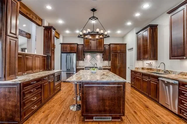 Before: the Lakewood Hills kitchen in Dallas, Texas, with dark wood cabinetry, a granite-topped island, hardwood floors, and a candle-style iron chandelier, prior to the Artbuckle & Co. renovation.