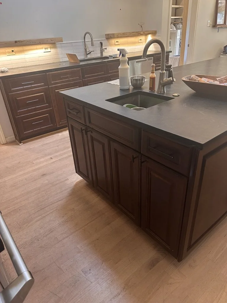 Before: the kitchen island in a Lakewood Hills home in Dallas, Texas, with dark raised-panel cabinetry and a black countertop with an undermount sink, prior to the Artbuckle & Co. renovation.