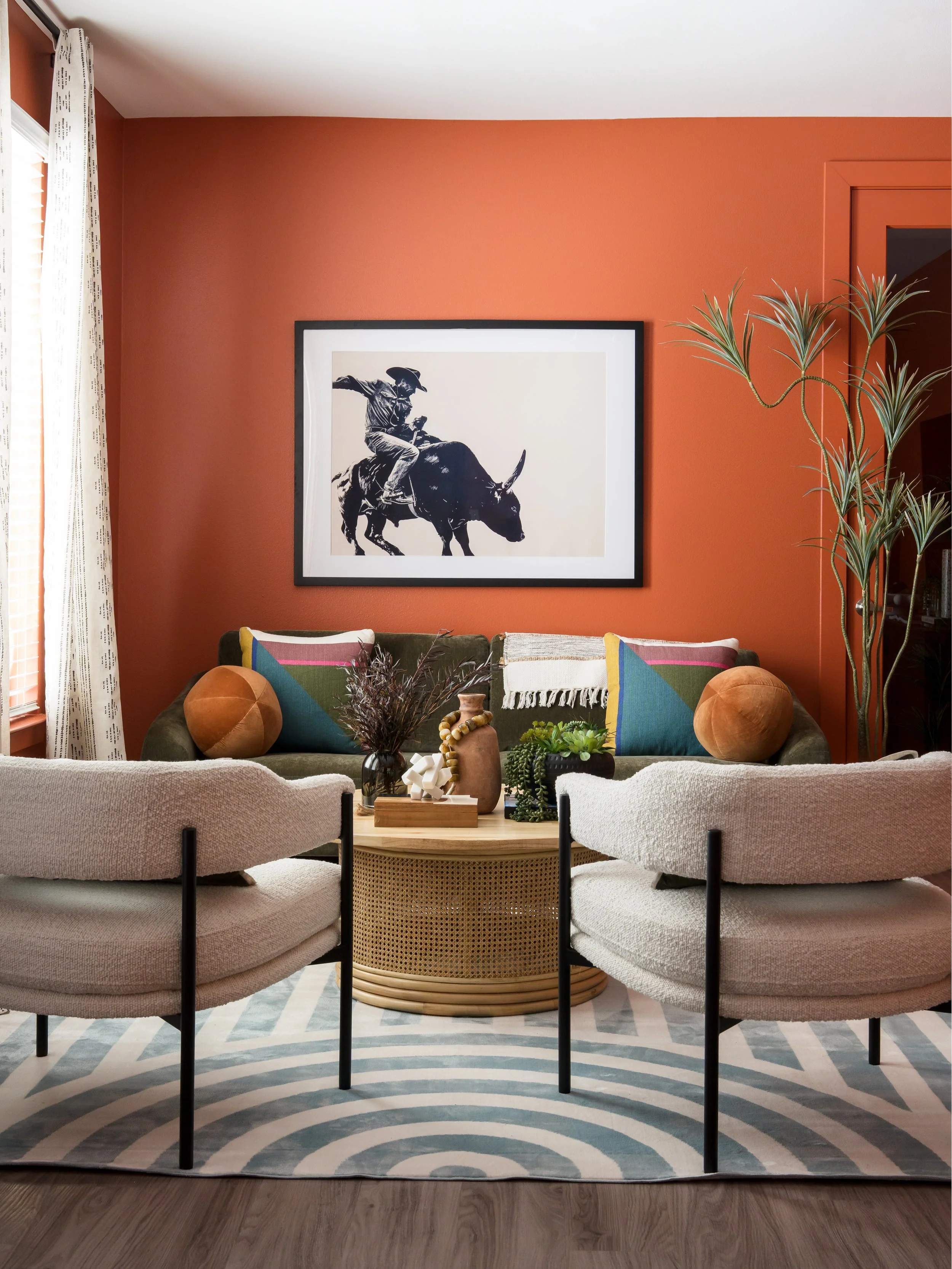 Living room with rust-colored wall, framed black-and-white artwork of a cowboy on a bull, cream-colored sofas with colorful pillows, a round wicker coffee table, plants, and patterned rug.