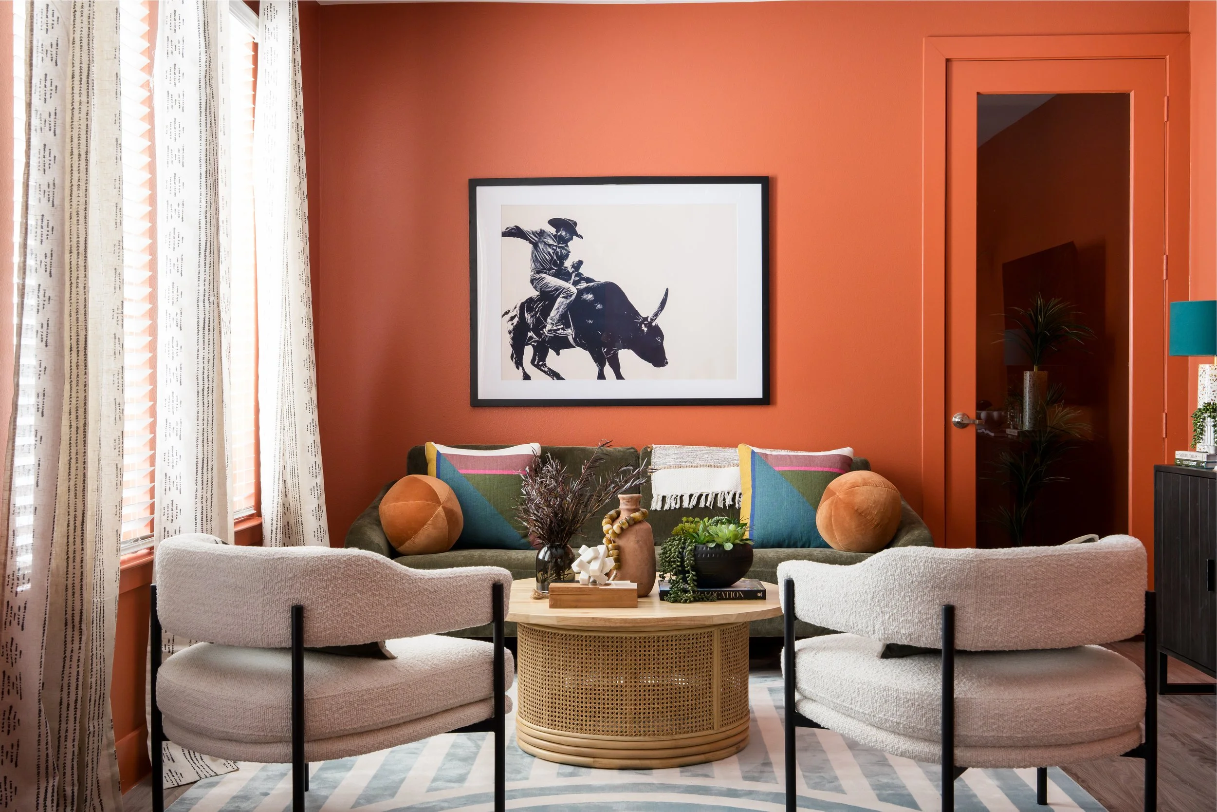 A cozy living room with a burnt orange wall, white curtains, and eclectic furniture including a cream-colored sofa with colorful pillows, a rattan coffee table with decorative items, and a large framed black and white art piece of a cowboy riding a b