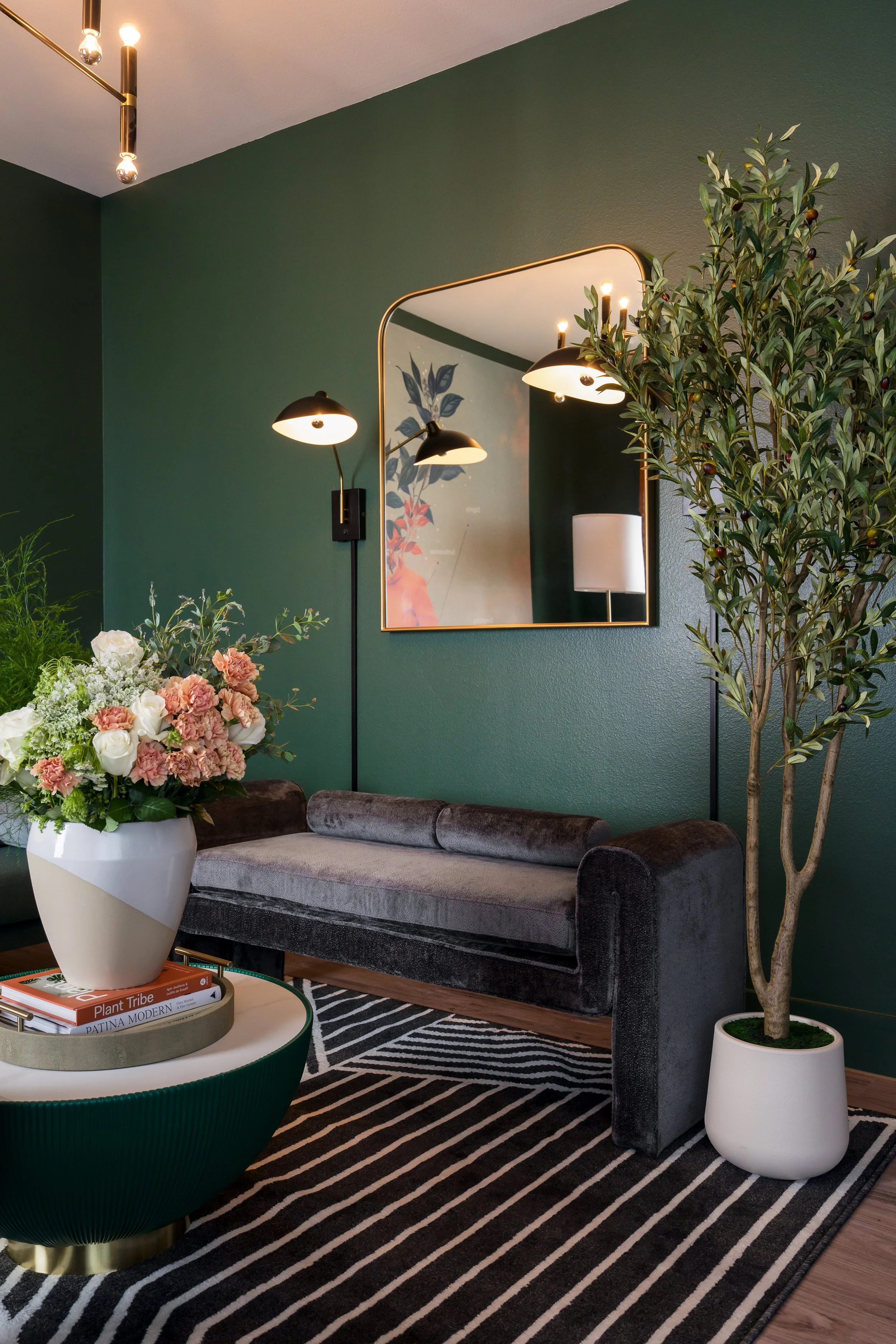 A living room with a green wall, a velvet gray sofa, a black and white striped rug, a tall potted olive tree, a round white coffee table with a green rim and books, a wall mirror, and wall-mounted black and gold lamps.