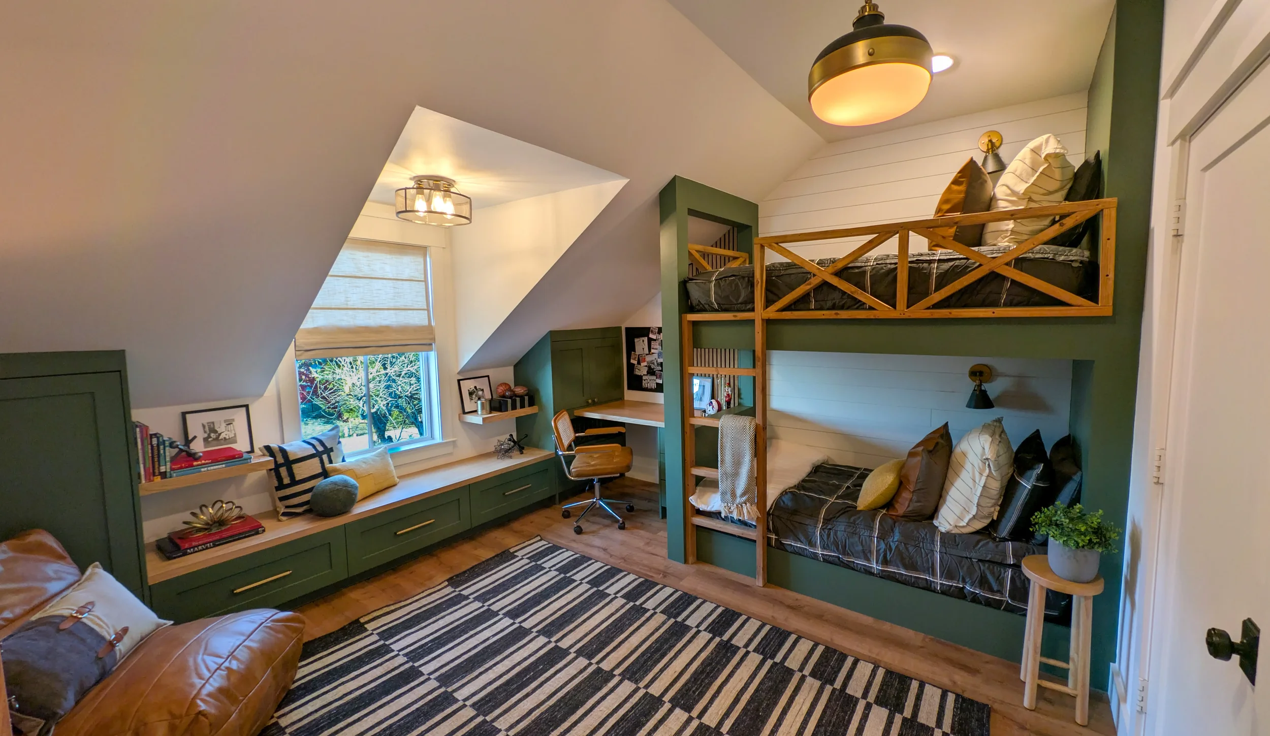 Room featuring a loft bed with black bedding and pillows, a window seat with decorative pillows, desk area with a chair, green cabinetry, and a striped rug on wooden floor. Artbuckle & Co.