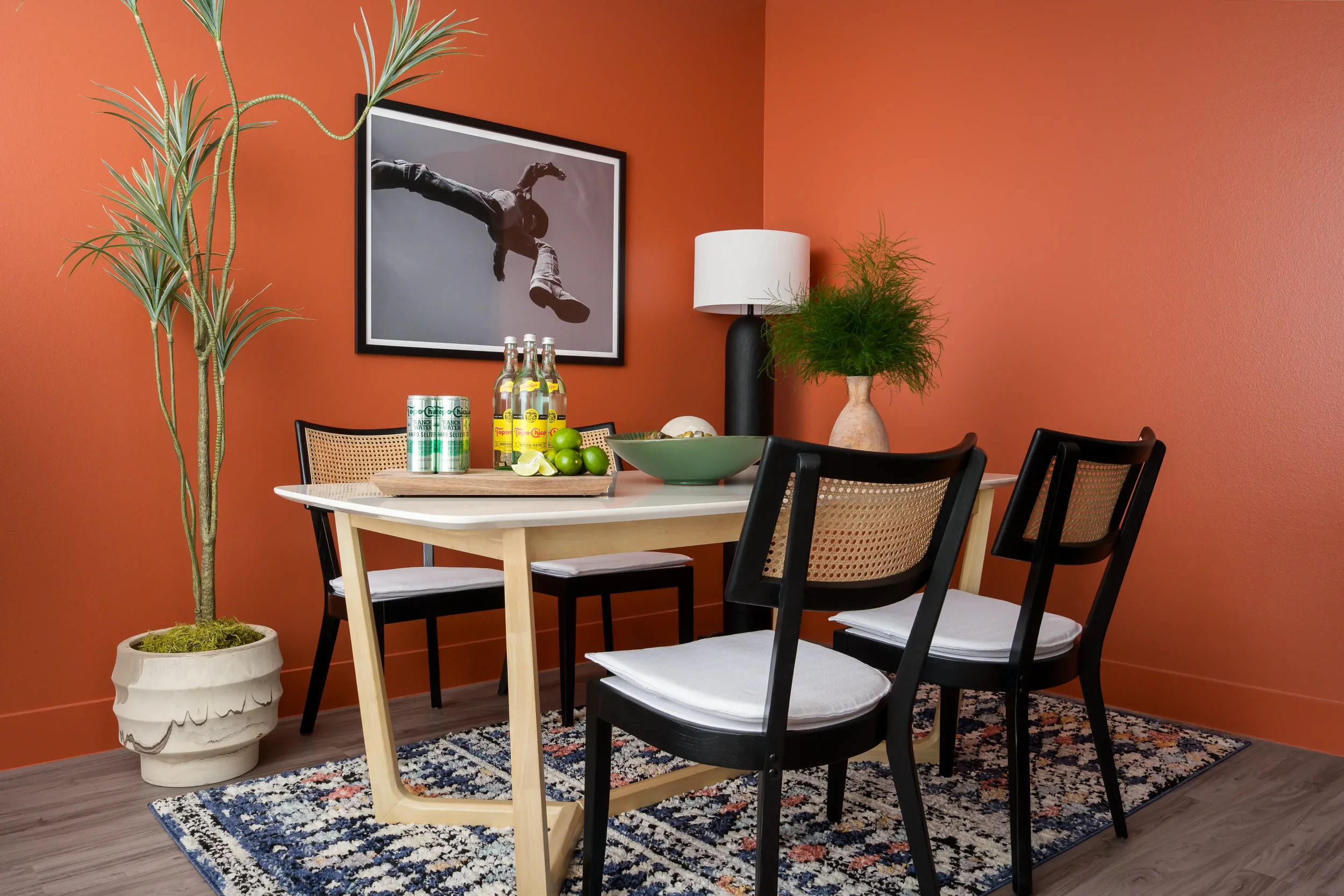 A dining room with an orange wall, a framed black and white photograph of a person performing a backflip, a white table with black chairs, a large potted plant, a table lamp, and a bowl of limes and lemons on the table.