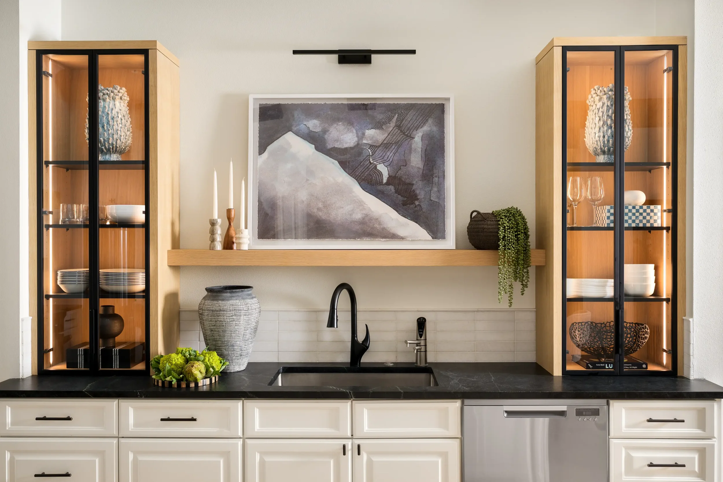 Modern kitchen with white cabinets, black countertop, and black sink. Two tall wooden display cabinets with glass doors contain decorative items. A large framed abstract artwork hangs on the wall above a wooden shelf, which holds three white candles and a woven basket with greenery. On the countertop, there are artichokes and leafy greens, and a large textured vase.