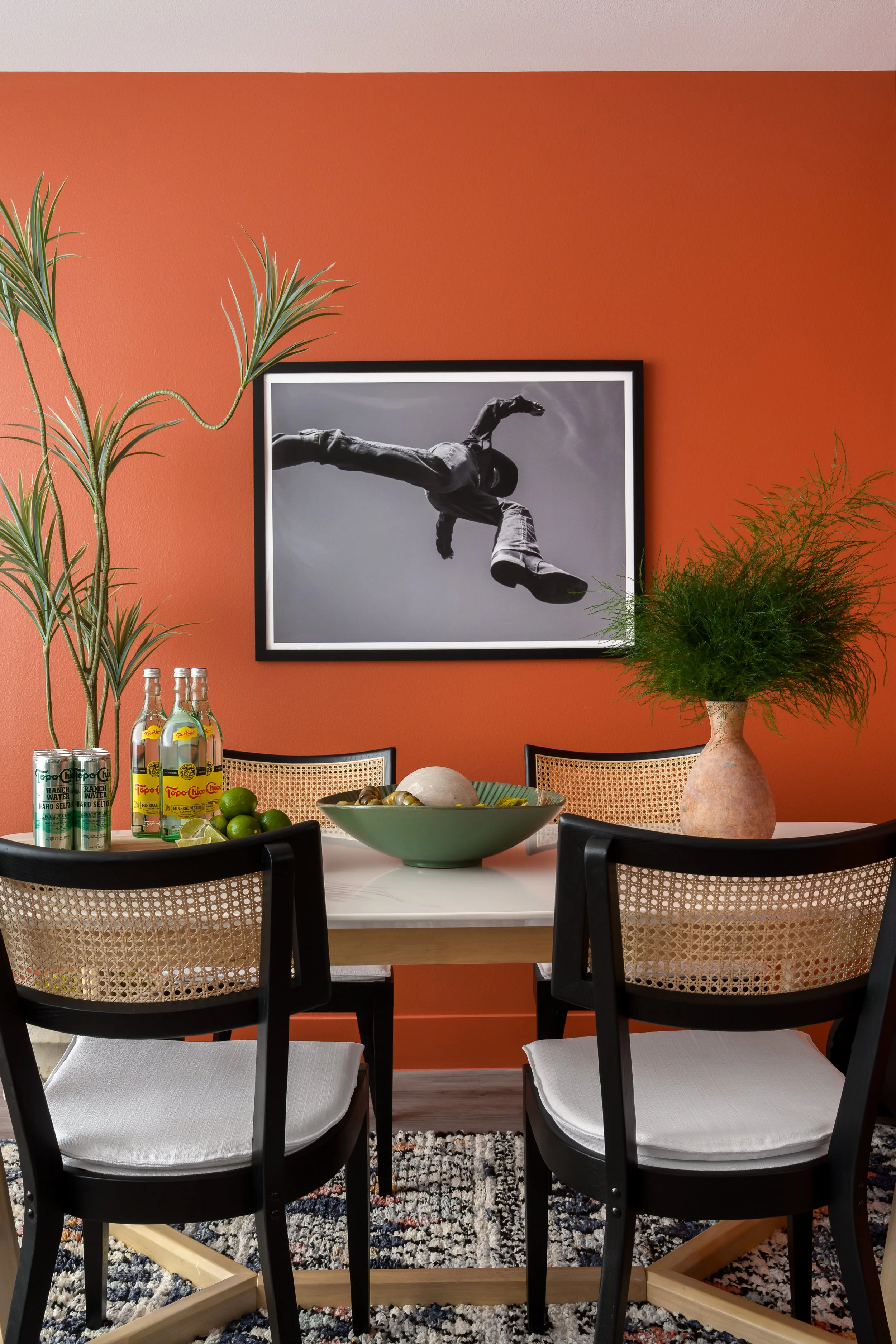 A dining room with a round table surrounded by four black chairs with white cushions. The table has a green bowl with fruit, a set of three bottles, and a bowl with snacks. A large potted plant and a framed black-and-white photograph of a person jump