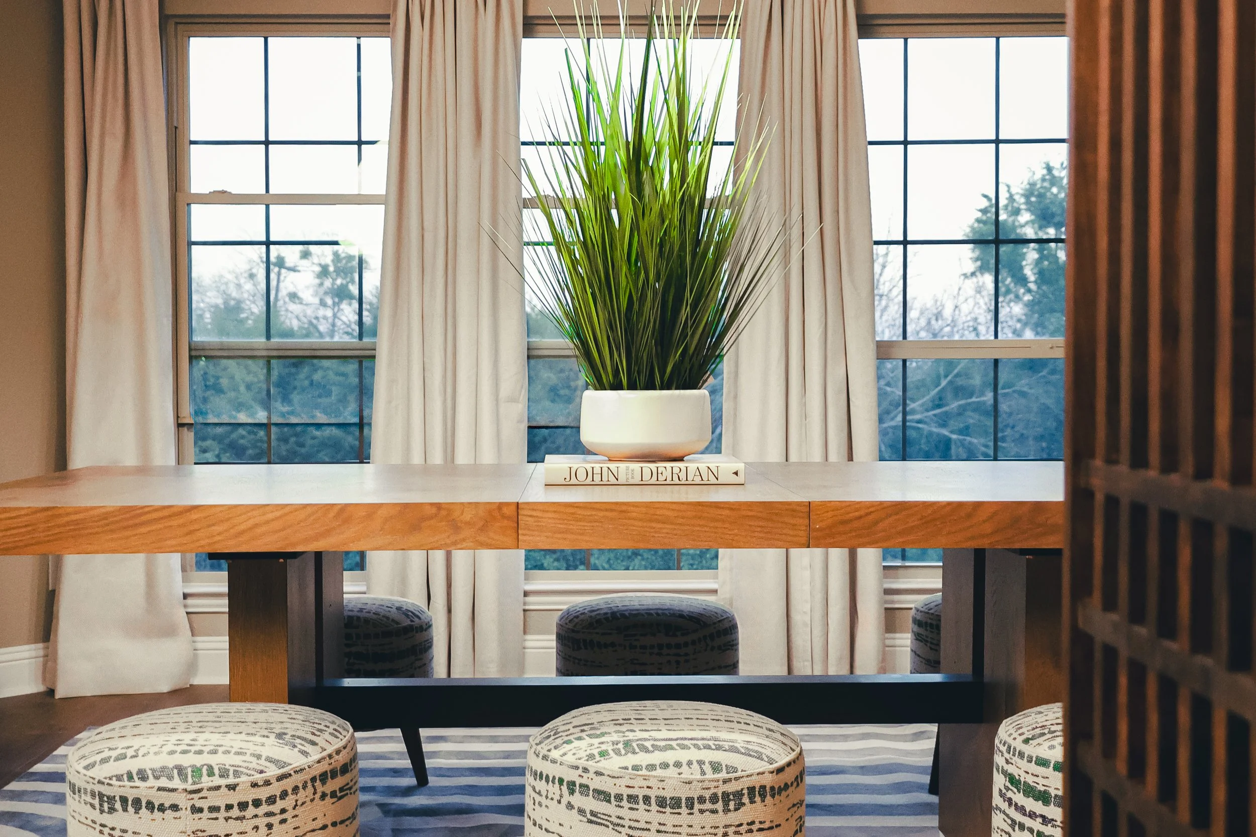 A window with white curtains framing the view of trees outside. A houseplant with tall green grass-like leaves in a white pot sits on a wooden table, which has a book underneath titled 'John Derian.' In front of the table, there are three patterned r