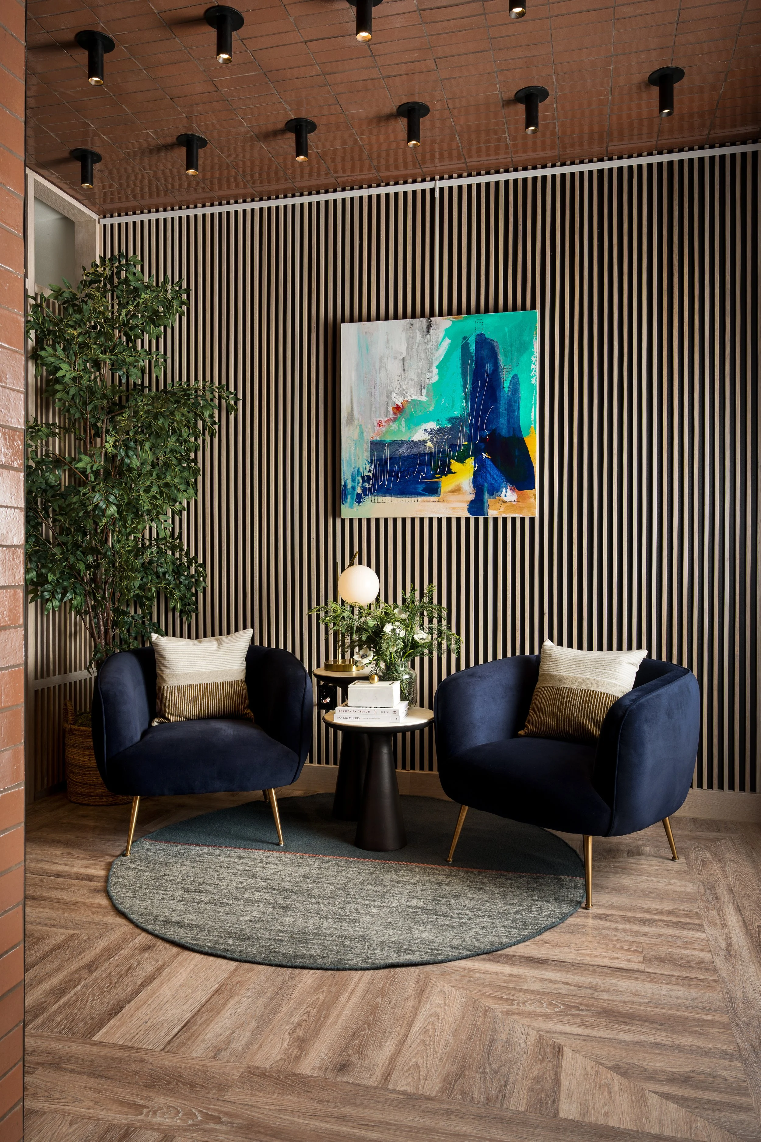 A cozy seating area with two navy blue velvet armchairs with gold legs, a small black round table with a lamp, books, and greenery, set against a wood-paneled wall with a colorful abstract painting, under a wooden ceiling with black track lighting an