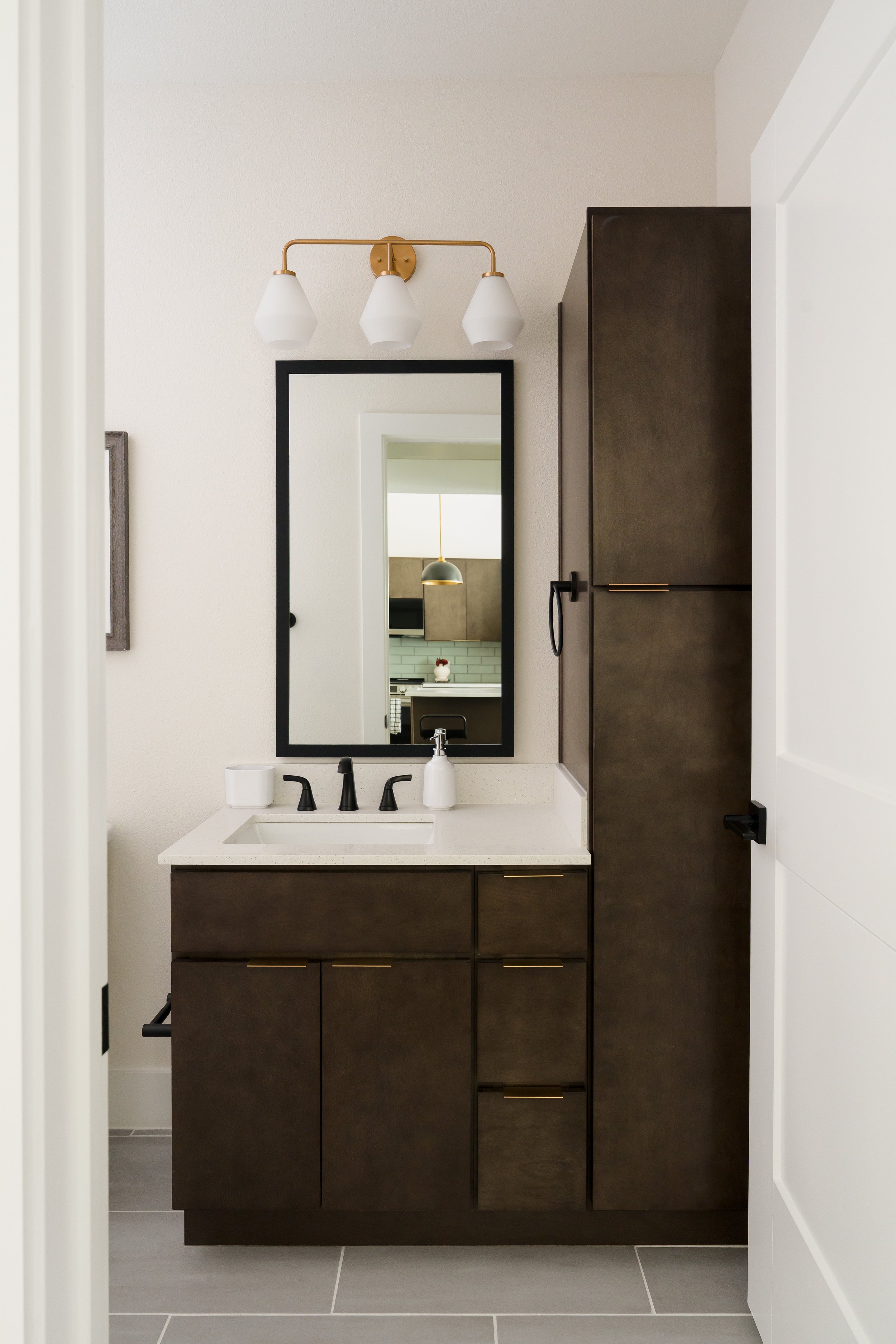 Apartment bathroom vanity at The Gallery in Frisco, Texas, featuring dark custom cabinetry, a brass 3-light sconce, matte black plumbing fixtures, and a black-framed mirror reflecting a ring pendant, designed by Artbuckle & Co.