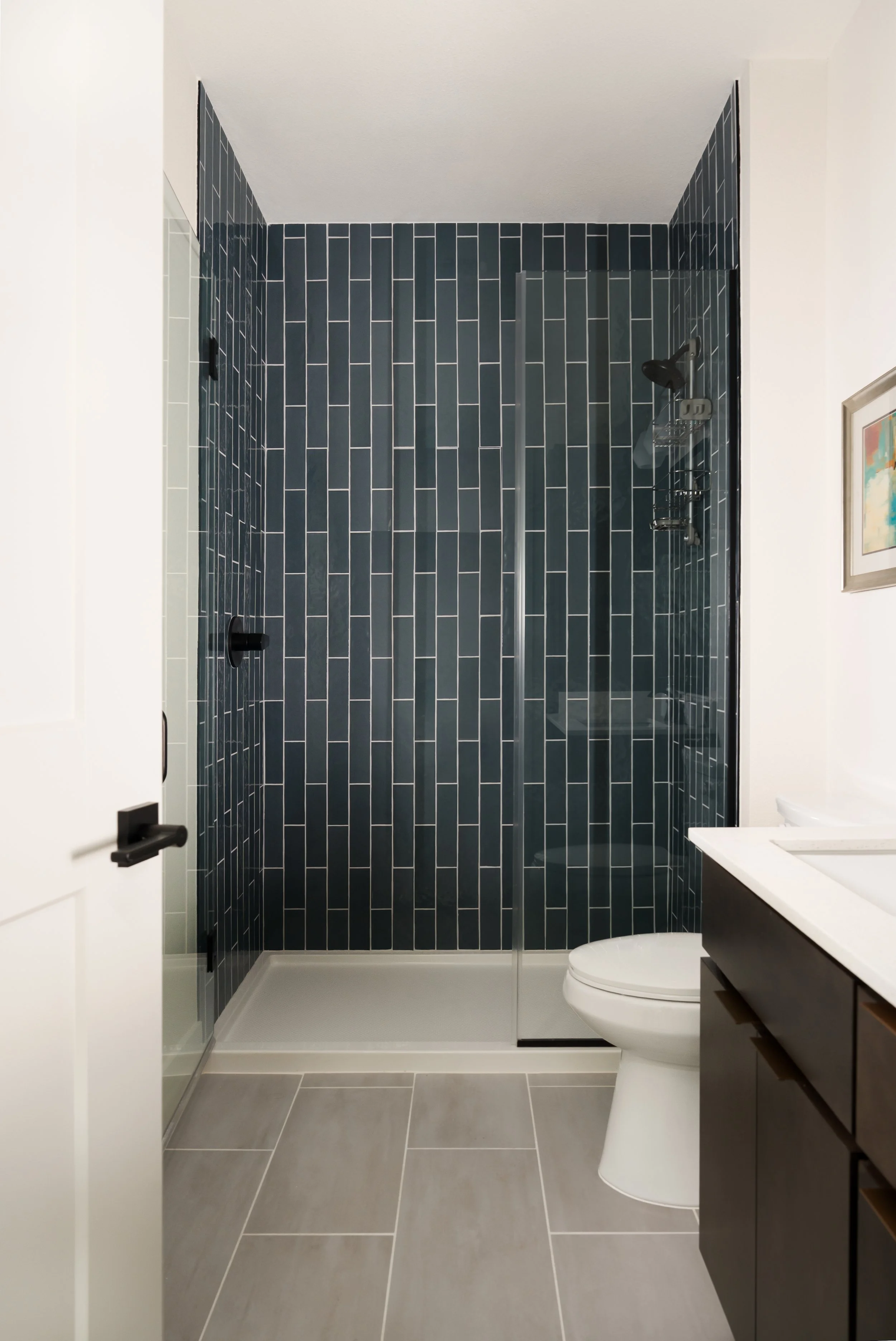 Apartment bathroom walk-in shower at The Gallery in Frisco, Texas, with vertical petrol blue subway tile, a glass enclosure, matte black fixtures, and light grey floor tile, designed by Artbuckle & Co.