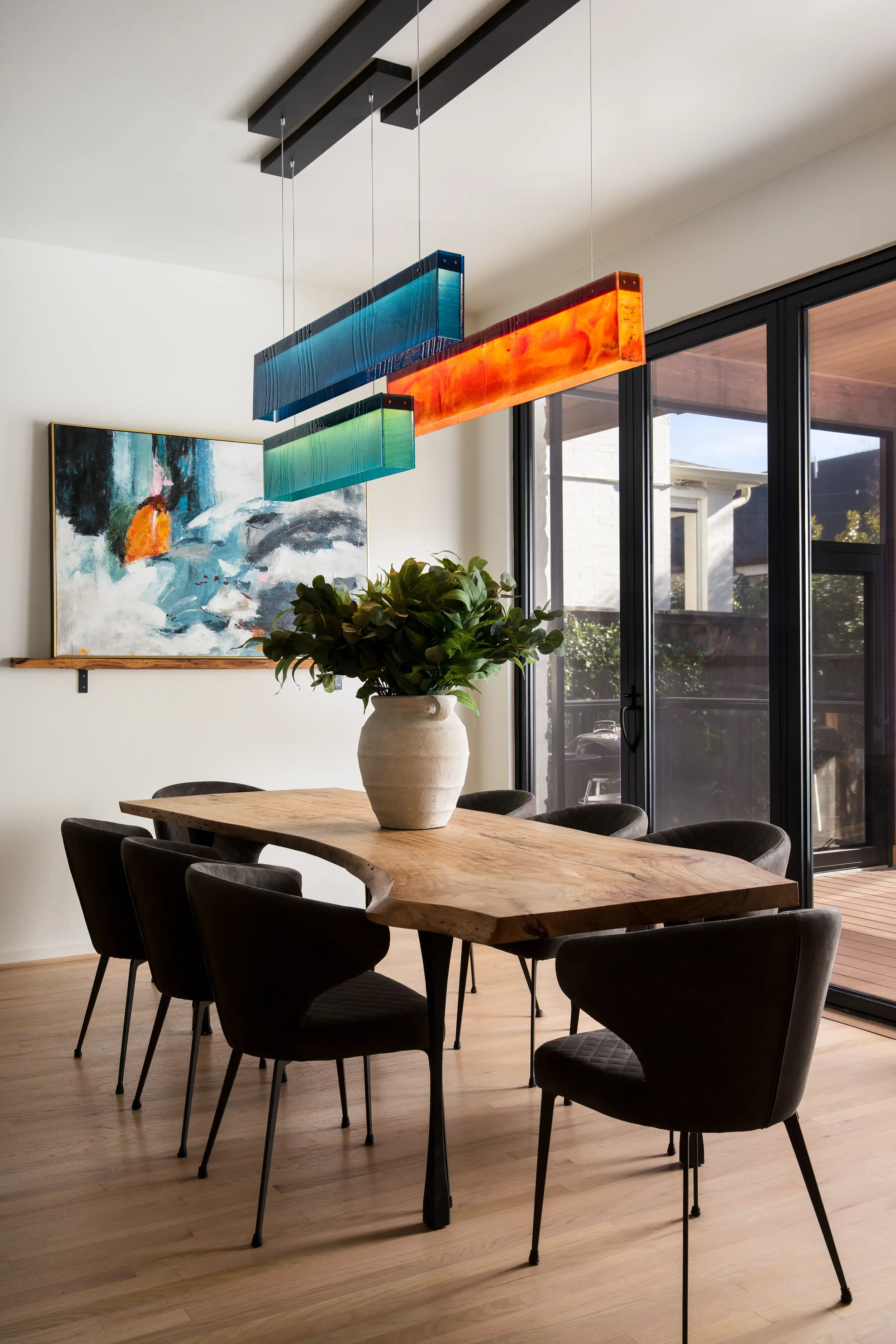 A dining room with a wooden table surrounded by black chairs. A large potted plant is centered on the table. There is modern, colorful hanging light fixtures above the table, and a large abstract painting on the wall. Large glass doors let in natural