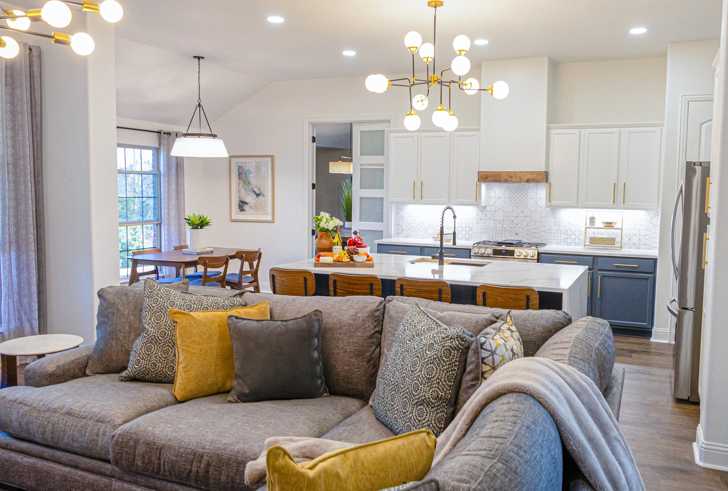 Open-concept living room and kitchen with modern lighting fixtures, patterned decor pillows on a large sectional sofa, dining table with chairs, and white cabinetry in the kitchen.