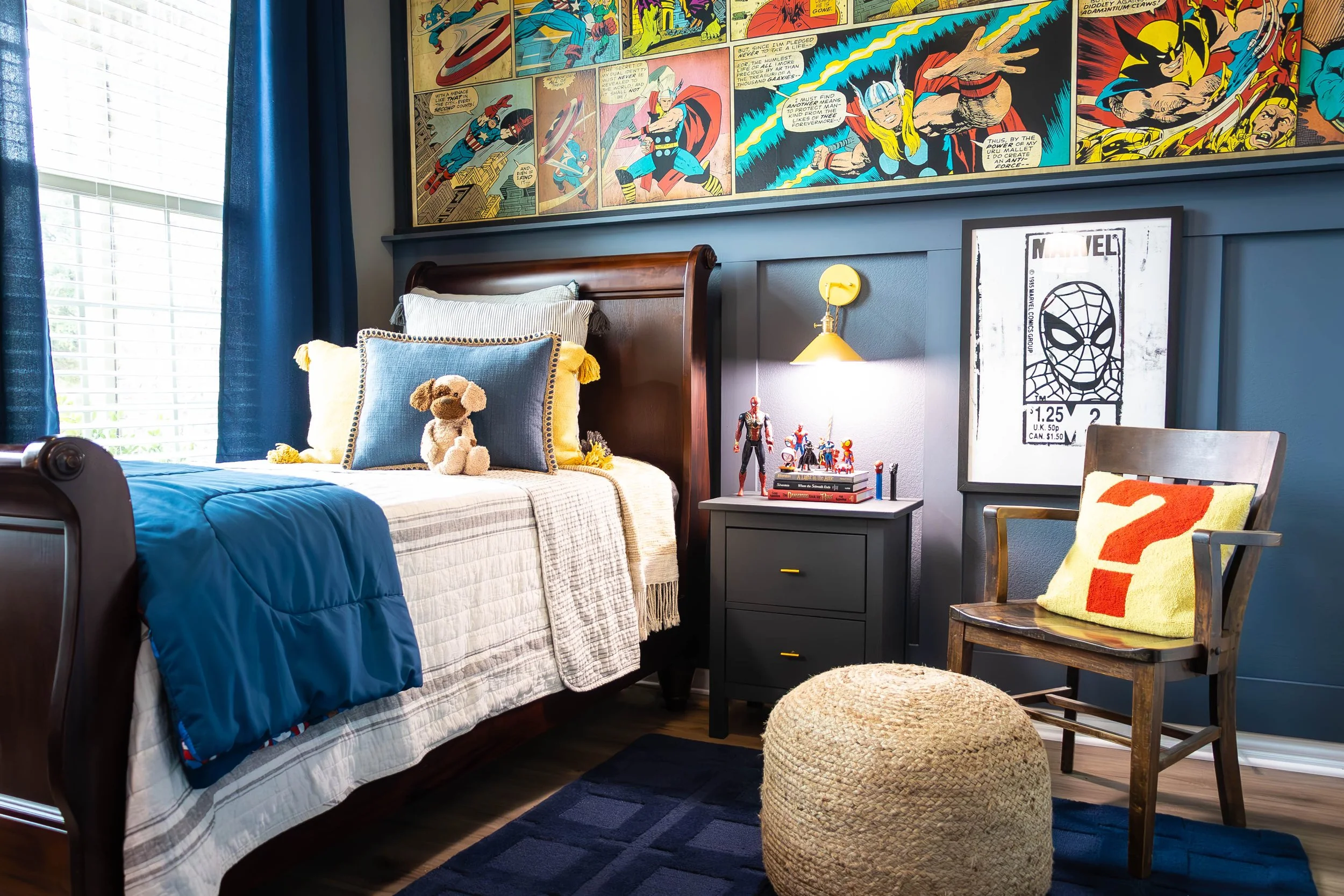 A bedroom with a wooden bed frame and blue bedding, a teddy bear on the pillows, and comic book wall art. There is a nightstand with superhero figurines, a lamp, and framed Marvel art. A wooden chair with a cushion and a woven pouf are in the room. B