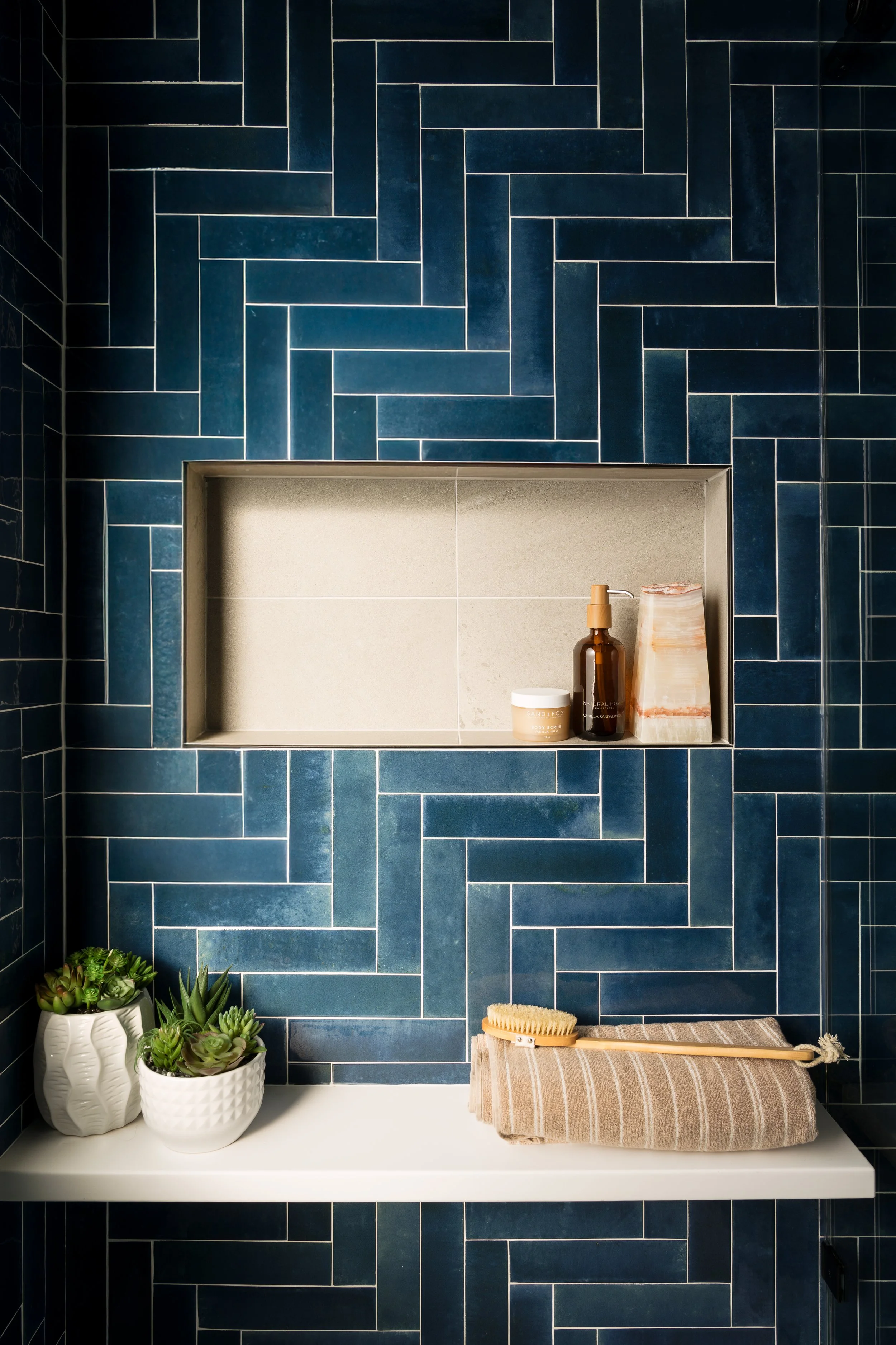 A bathroom shelf with a beige surface and a dark blue tile wall in the background. On the shelf, there are three potted succulents, a brown bottle with a pump, a smaller jar, a rectangular stone, a hairbrush, and a rolled towel.