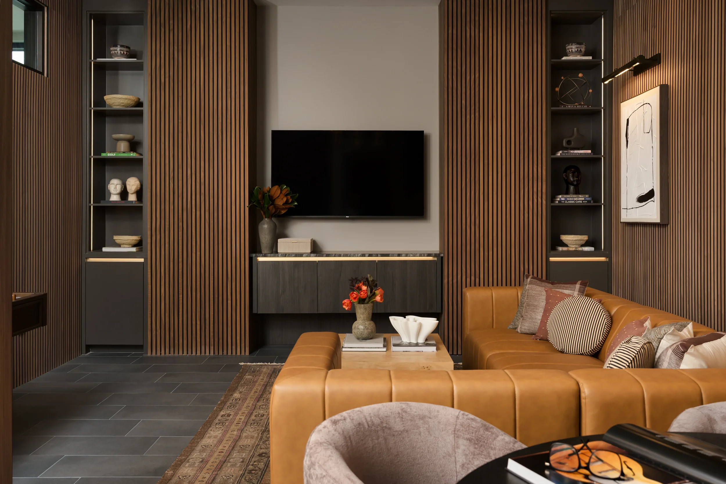 Media wall at The Platform in Frisco, Texas, with slatted walnut panels flanking a wall-mounted TV, a caramel leather sectional, and a layered rug, designed by Artbuckle & Co.