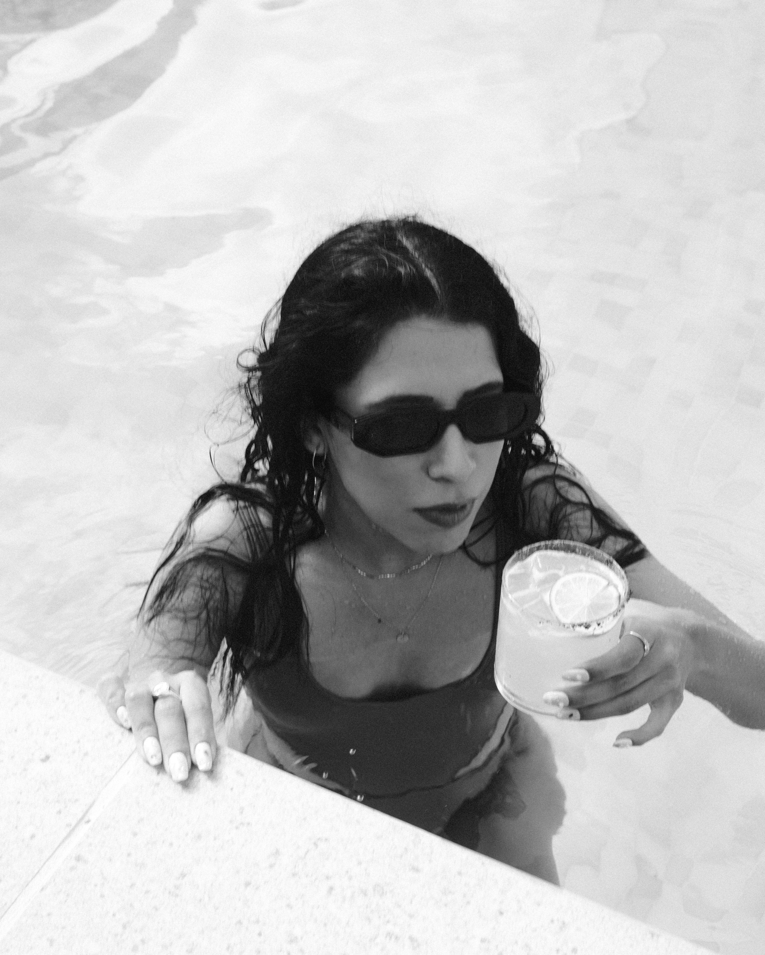 A woman with curly dark hair wearing sunglasses, holding a lemon cocktail with a slice of lemon, in a swimming pool.