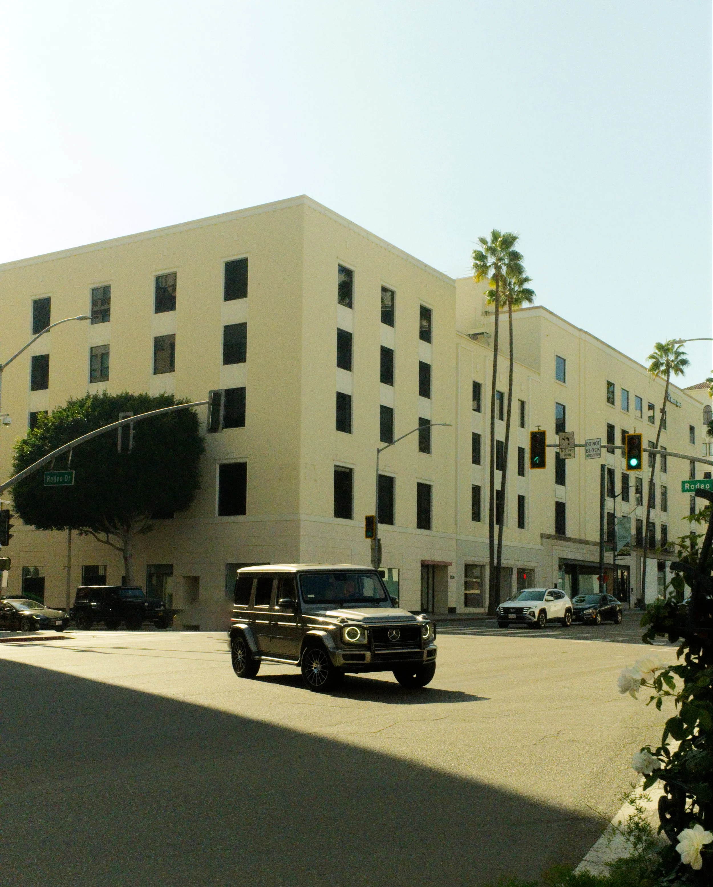 Street view showing a white multi-story building with multiple windows, palm trees, traffic lights, and a few cars including a black Mercedes-Benz G-Wagon, on Rodeo Drive.