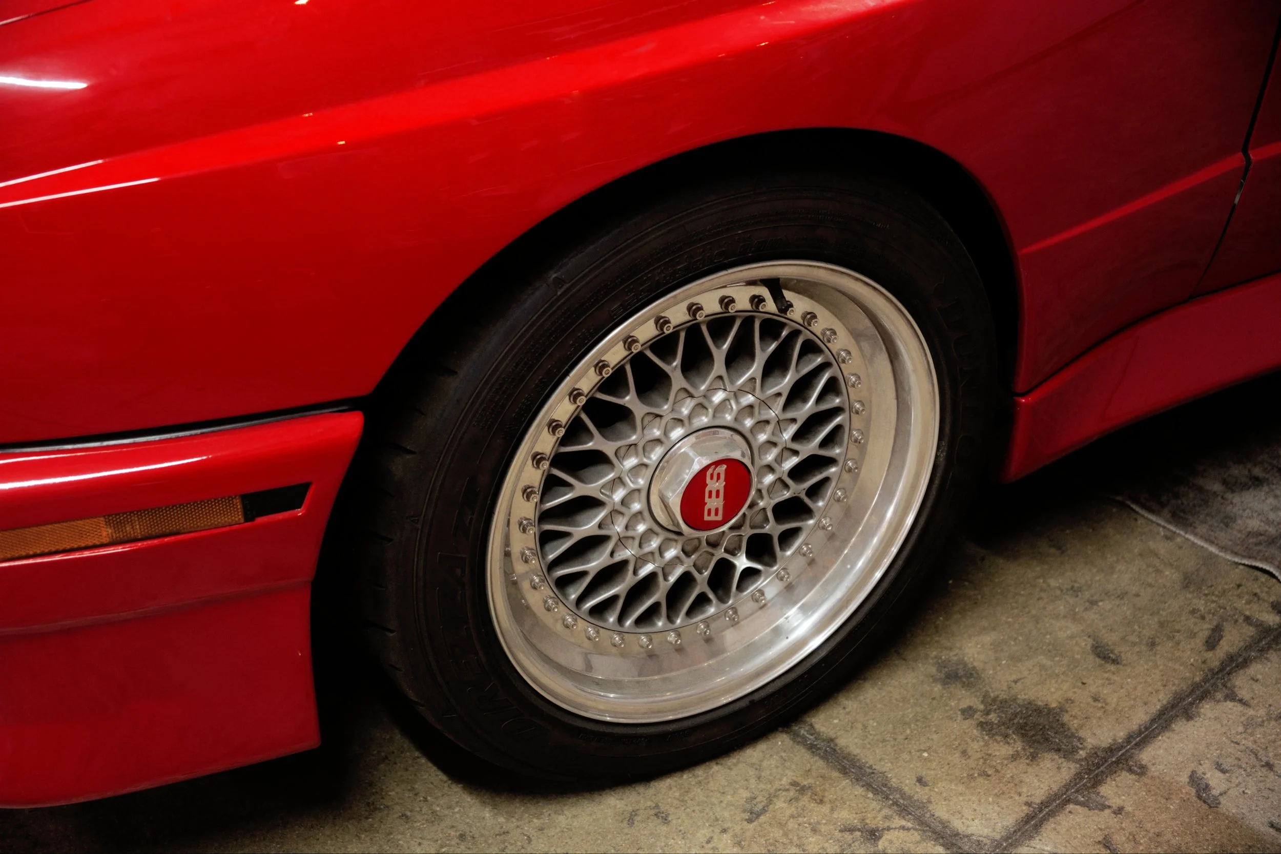 Close-up of a red sports car's front wheel with a silver BBS alloy rim and black tire, parked on concrete floor.