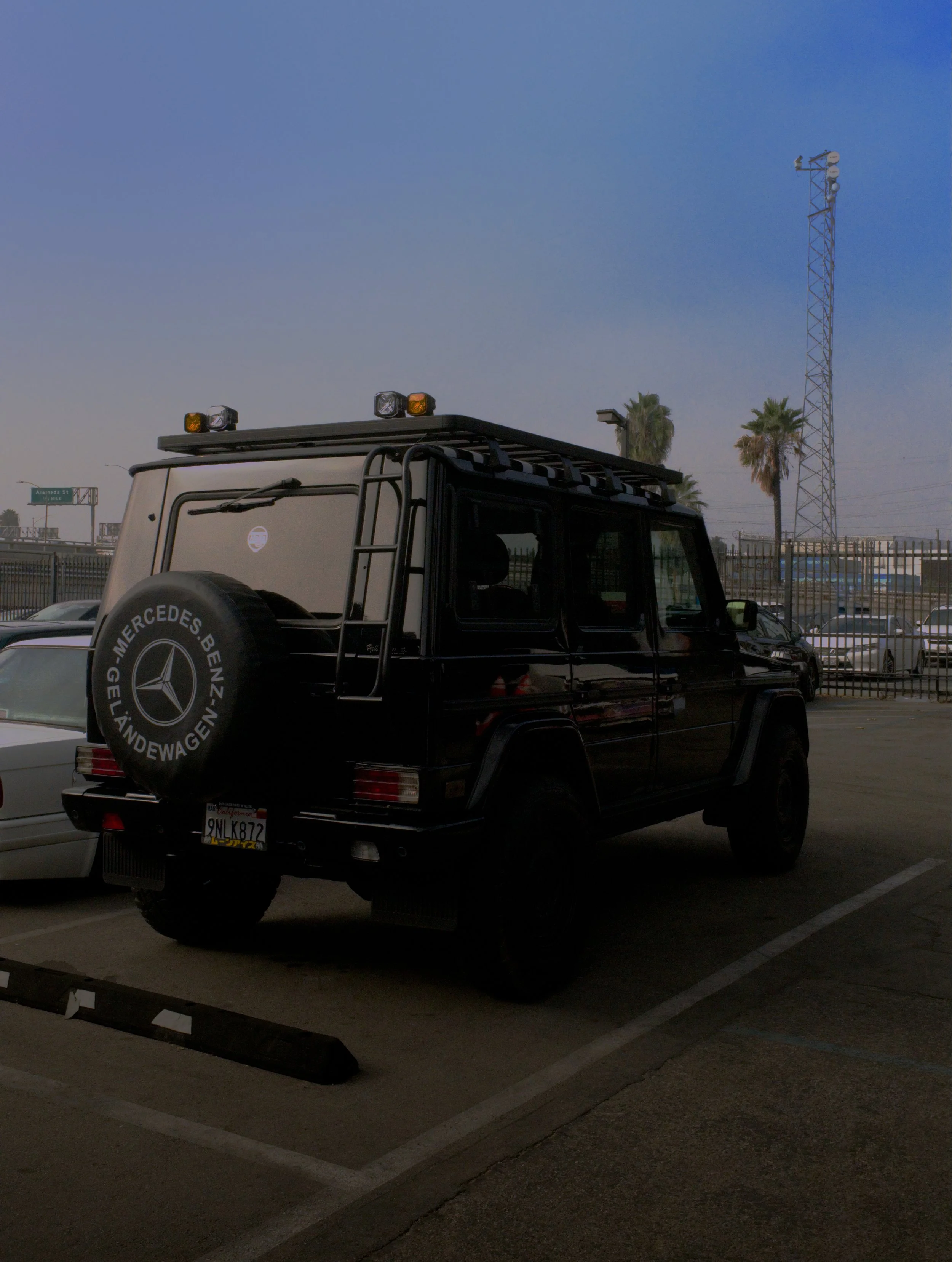 A black Mercedes-Benz G-Class SUV parked in a lot with palm trees in the background, a spare tire on the back with Mercedes branding, and a clear blue sky.