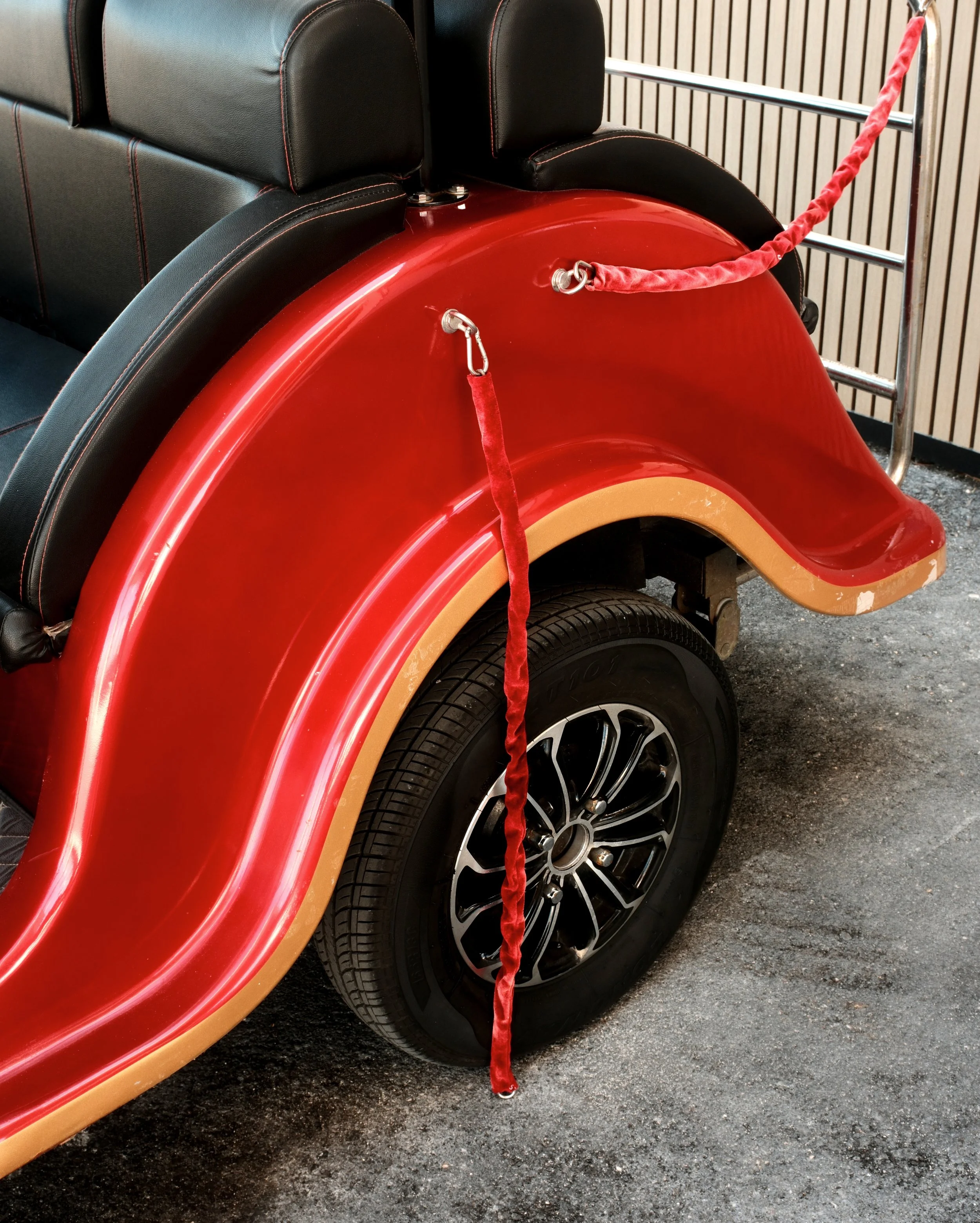 The rear wheel and fender of a small electric golf cart with a red and tan color scheme, black seats, and a red velvet rope barrier.