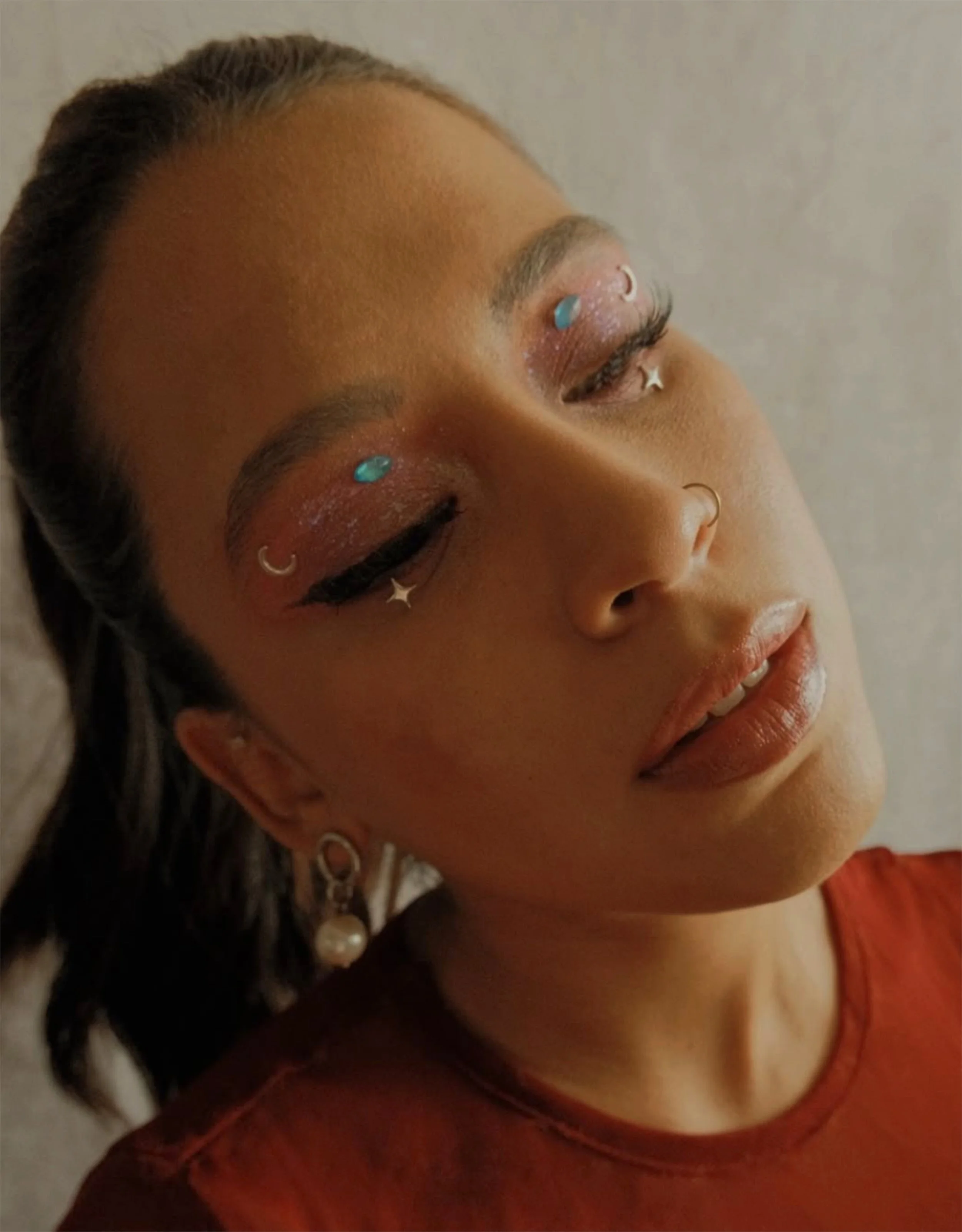 A woman with makeup featuring glitter eyeshadow, glitter accents, turquoise gemstones above her eyebrows, and star-shaped decorations near her eyes, wearing pearl earrings and a red top, with her eyes closed.