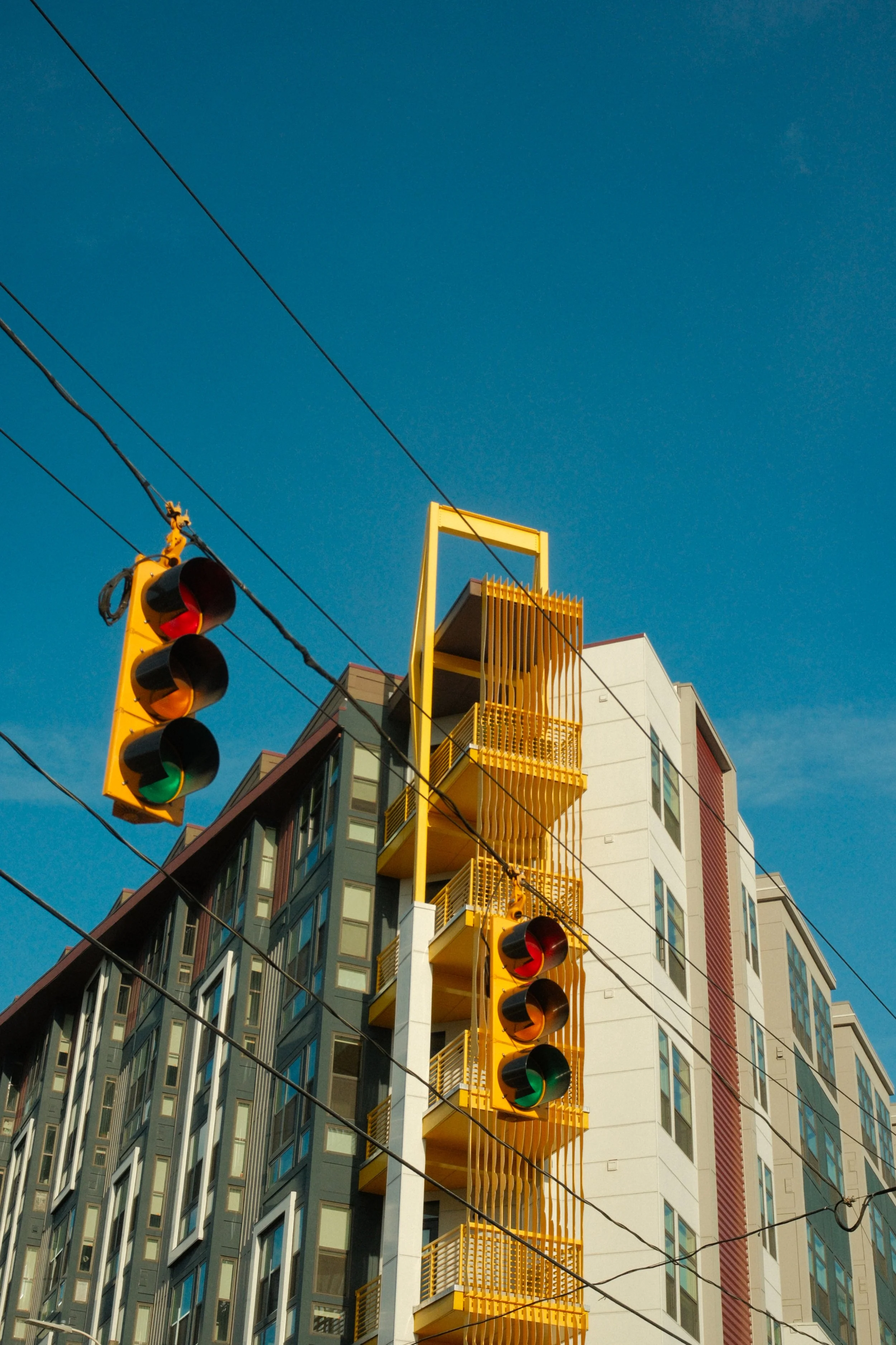 A multi-story building with a yellow fire escape and yellow framing around the stairwell, under a blue sky. Traffic lights are visible in the foreground.