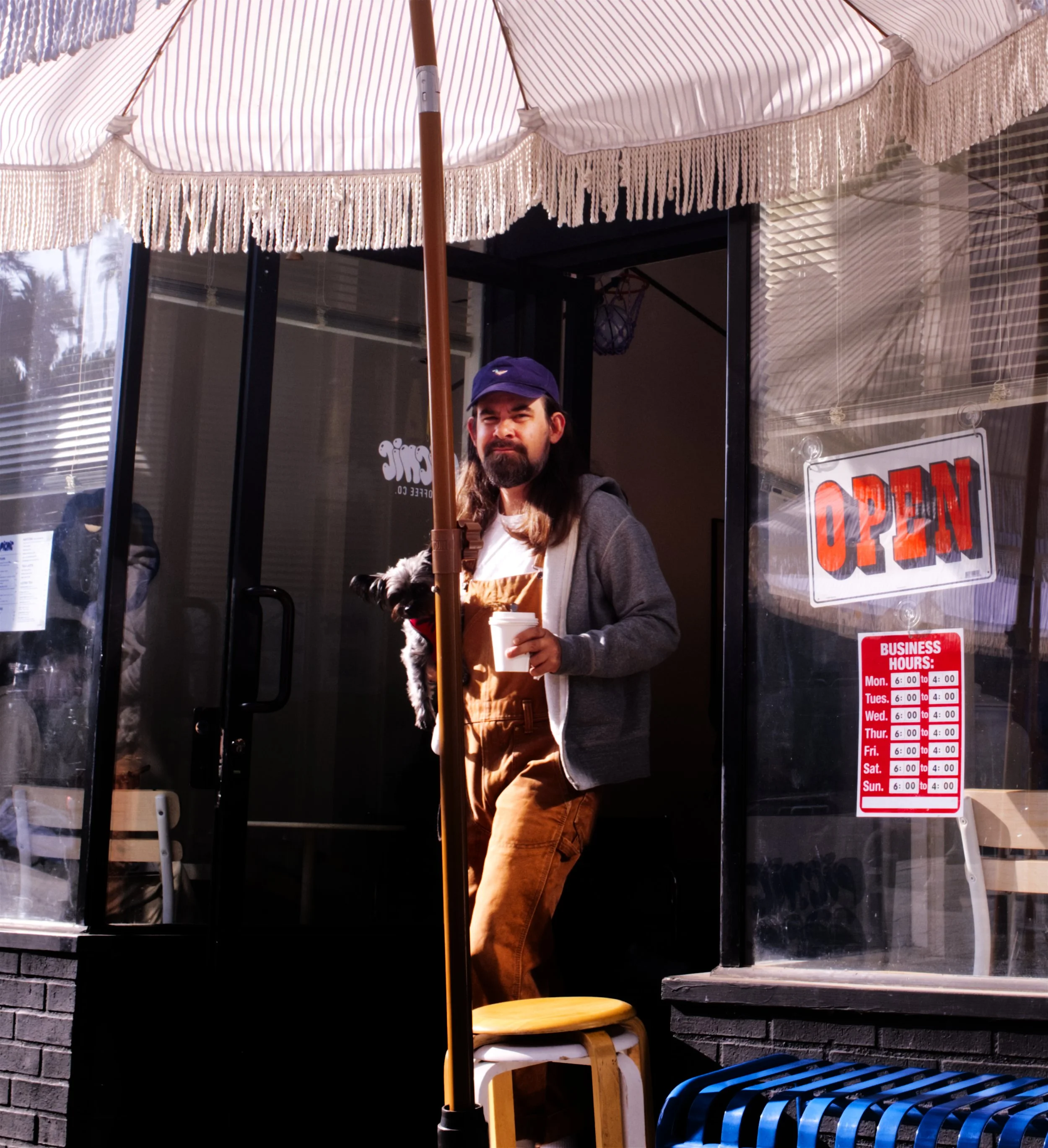 A man standing outside a cafe or restaurant with an 'OPEN' sign on the door, holding a coffee cup and a small dog. The cafe has a striped umbrella and business hours posted on the window.