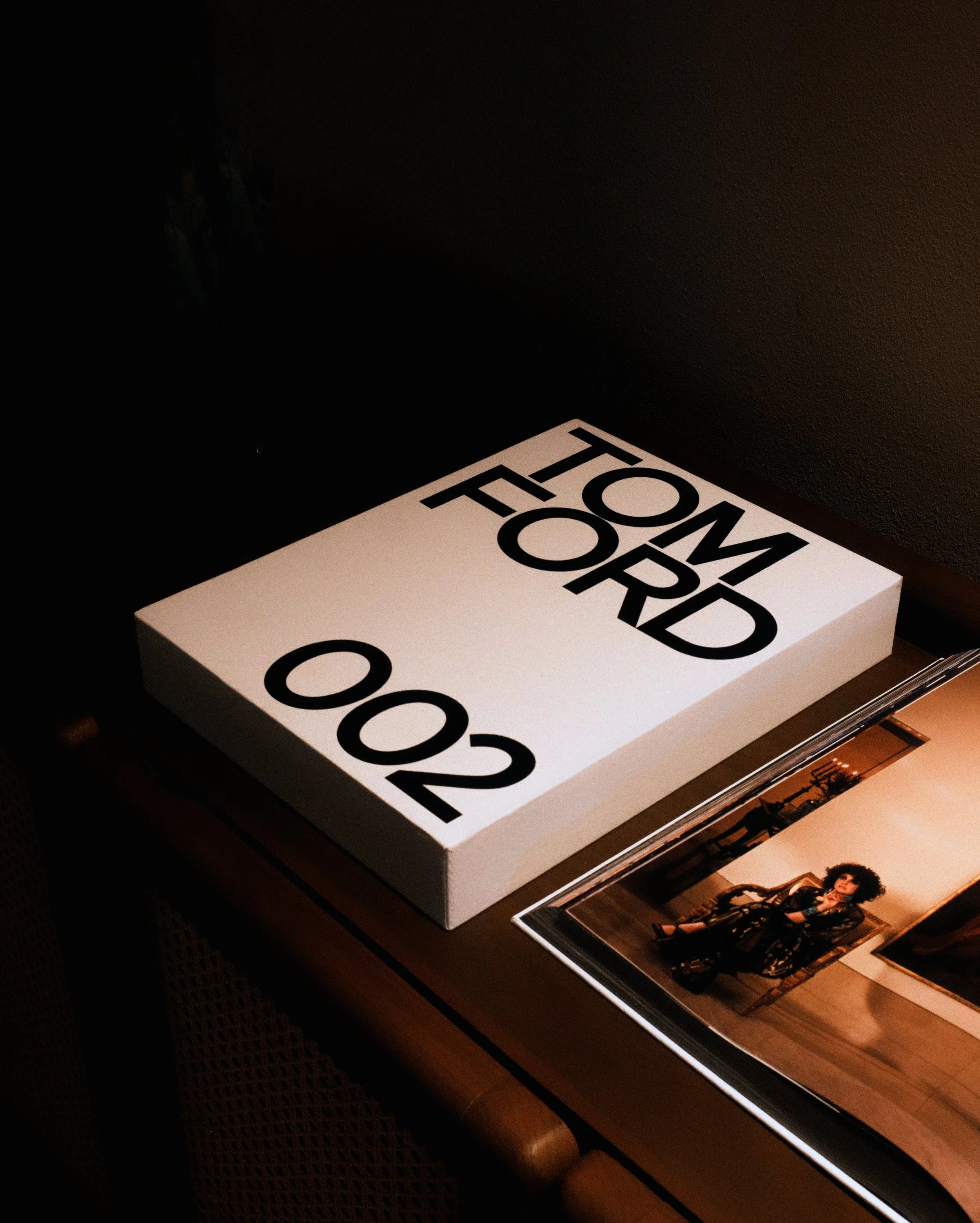 A white box with black text that reads "TOM FORD 002" on top, resting on a dark surface next to a photo album with pictures of a woman.