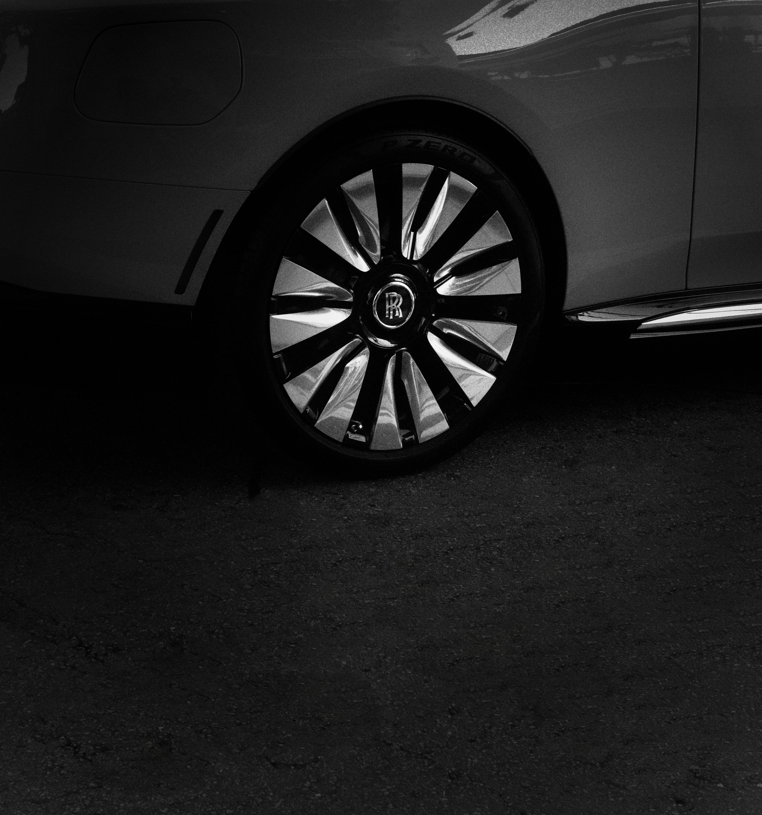 Close-up image of a luxury car's front wheel with a chrome multi-spoke rim and a Rolls-Royce logo in the center, shown in black and white.
