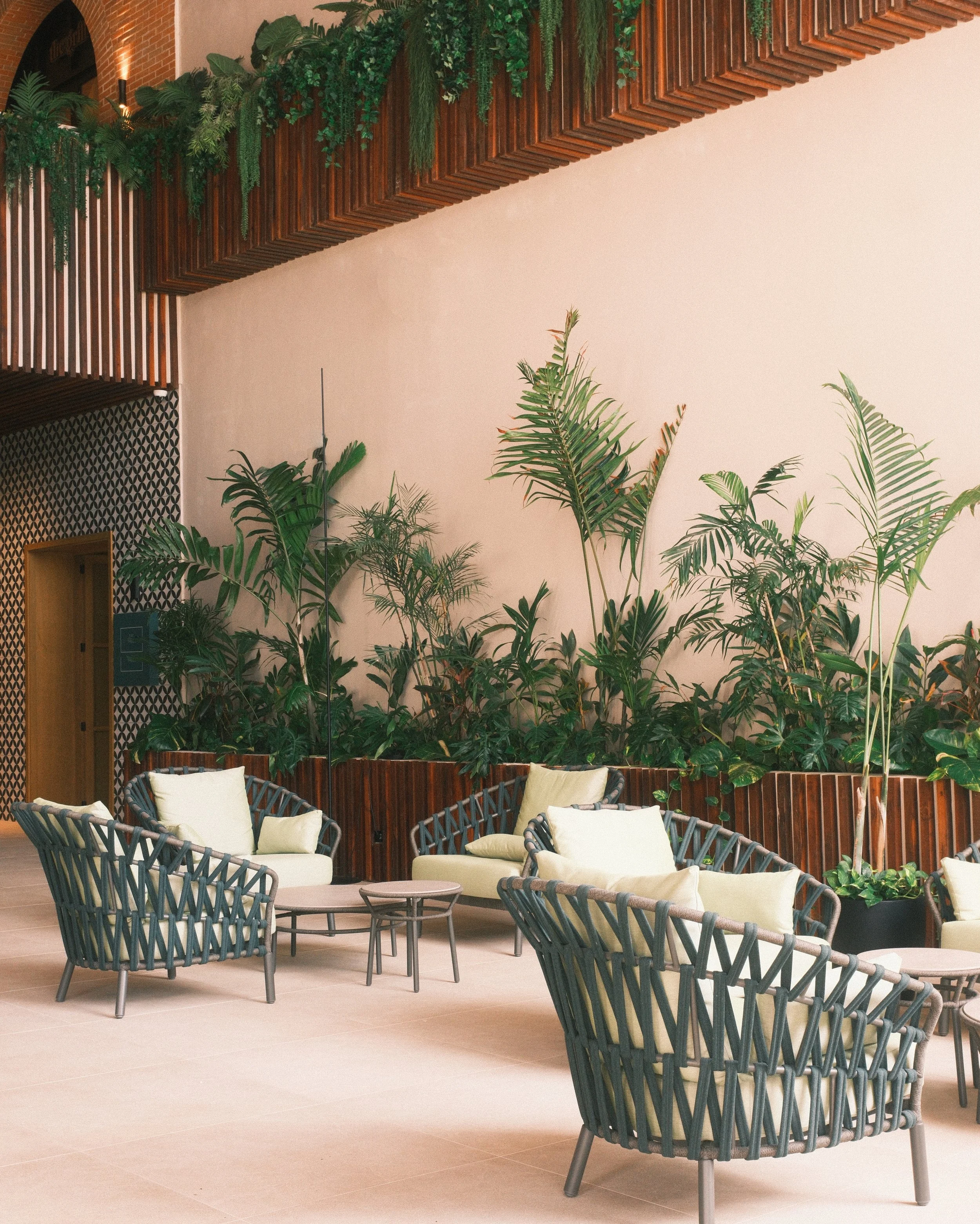 A cozy hotel lobby area with four modern round lounge chairs with cushions, small round tables, and lush green indoor plants against a beige wall, featuring wooden and patterned decor accents.