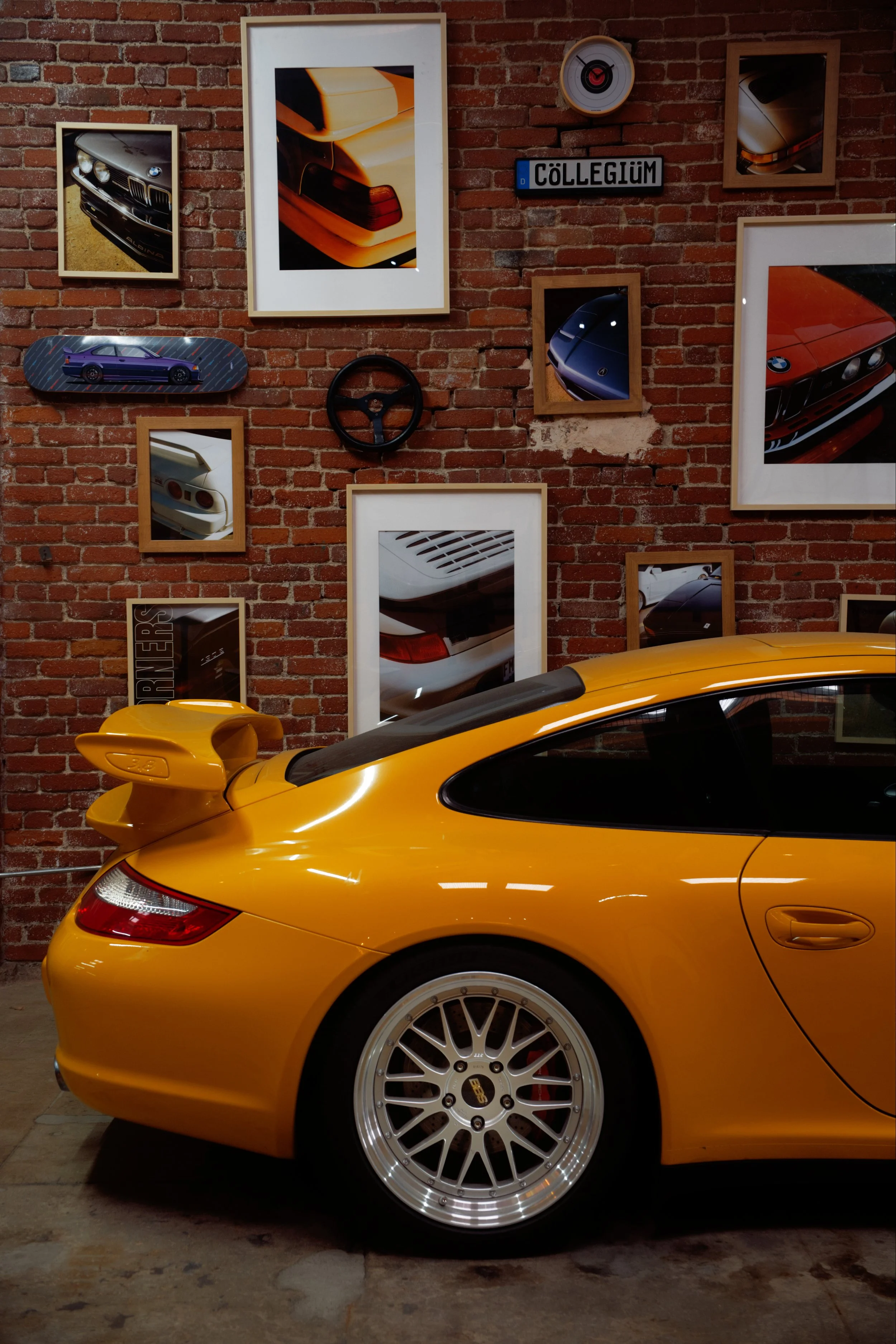A yellow Porsche sports car with a large rear wing is parked inside a garage with an exposed brick wall. The wall features multiple framed photos of cars, a circular wall clock, a vintage license plate that reads "CÖLLEGIUM," and a skateboard. The ca