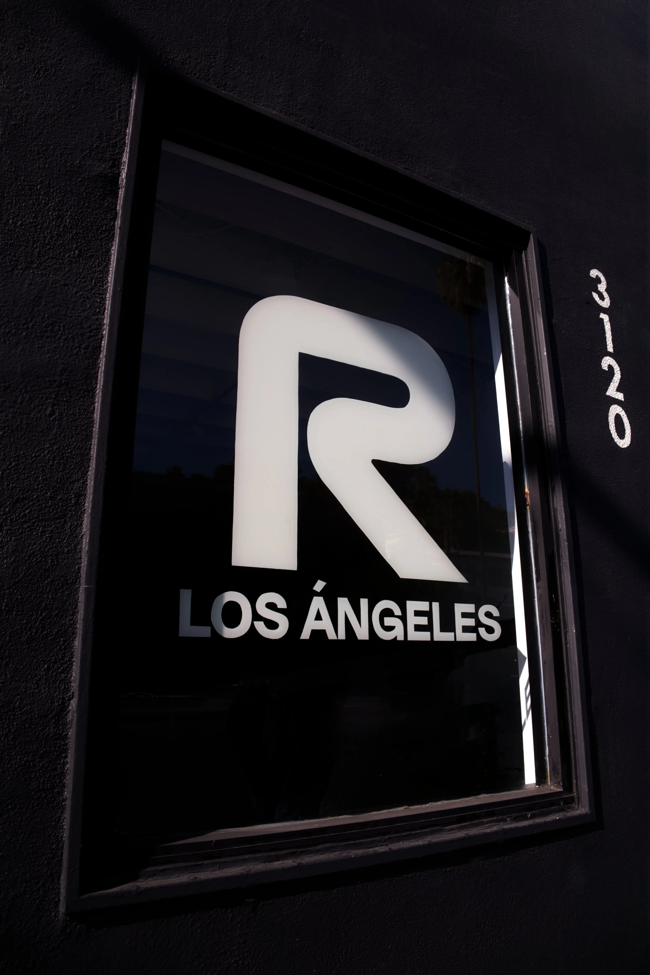 Storefront window with a white illuminated logo of the letter 'R' and the text 'LOS ÁNGELES' underneath, on a black wall with the address '3-20' written on the wall beside it.