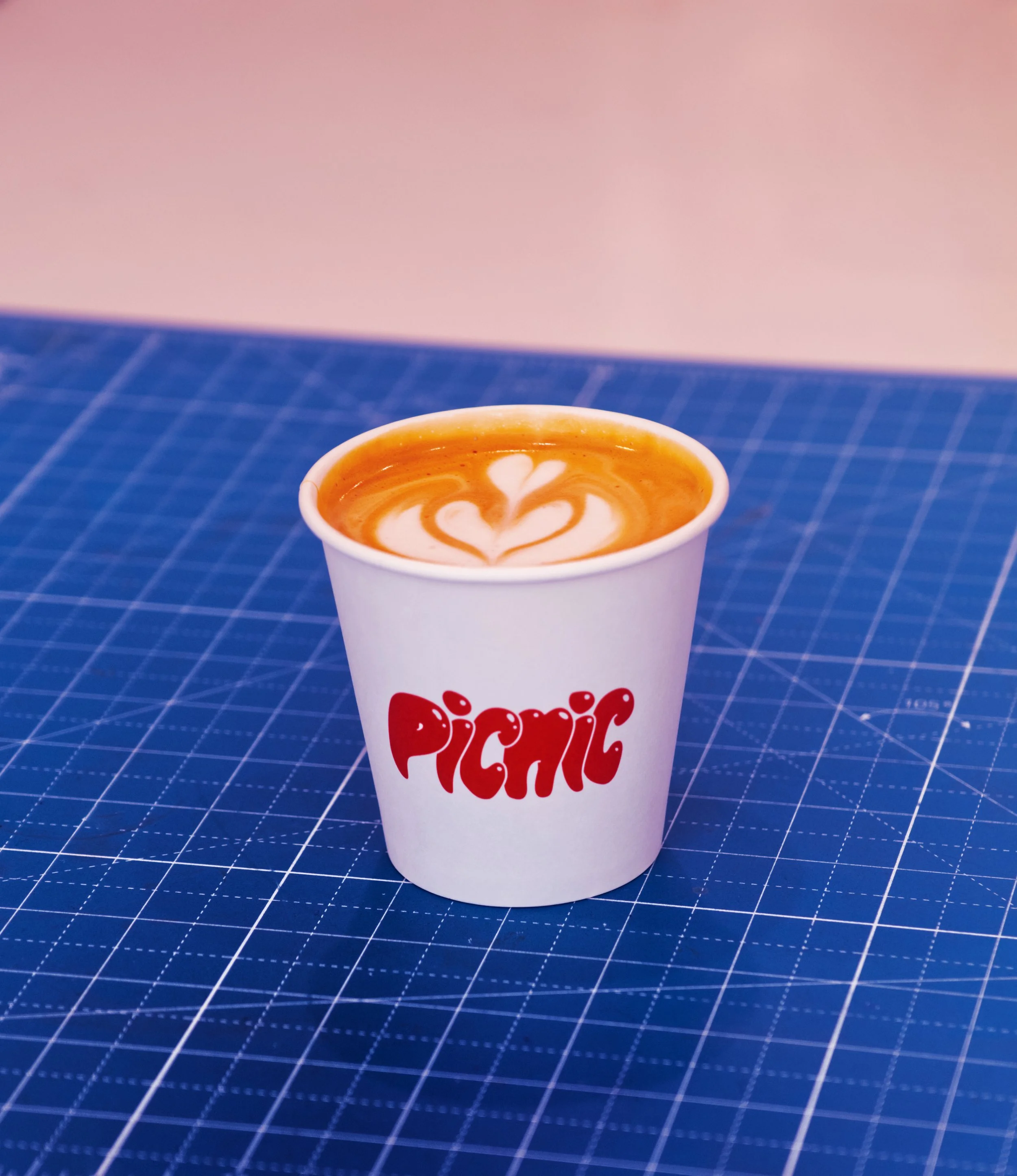 A paper cup with the word 'PICNIC' written in red, containing a latte with heart and leaf-shaped foam art, placed on a blue grid cutting mat.