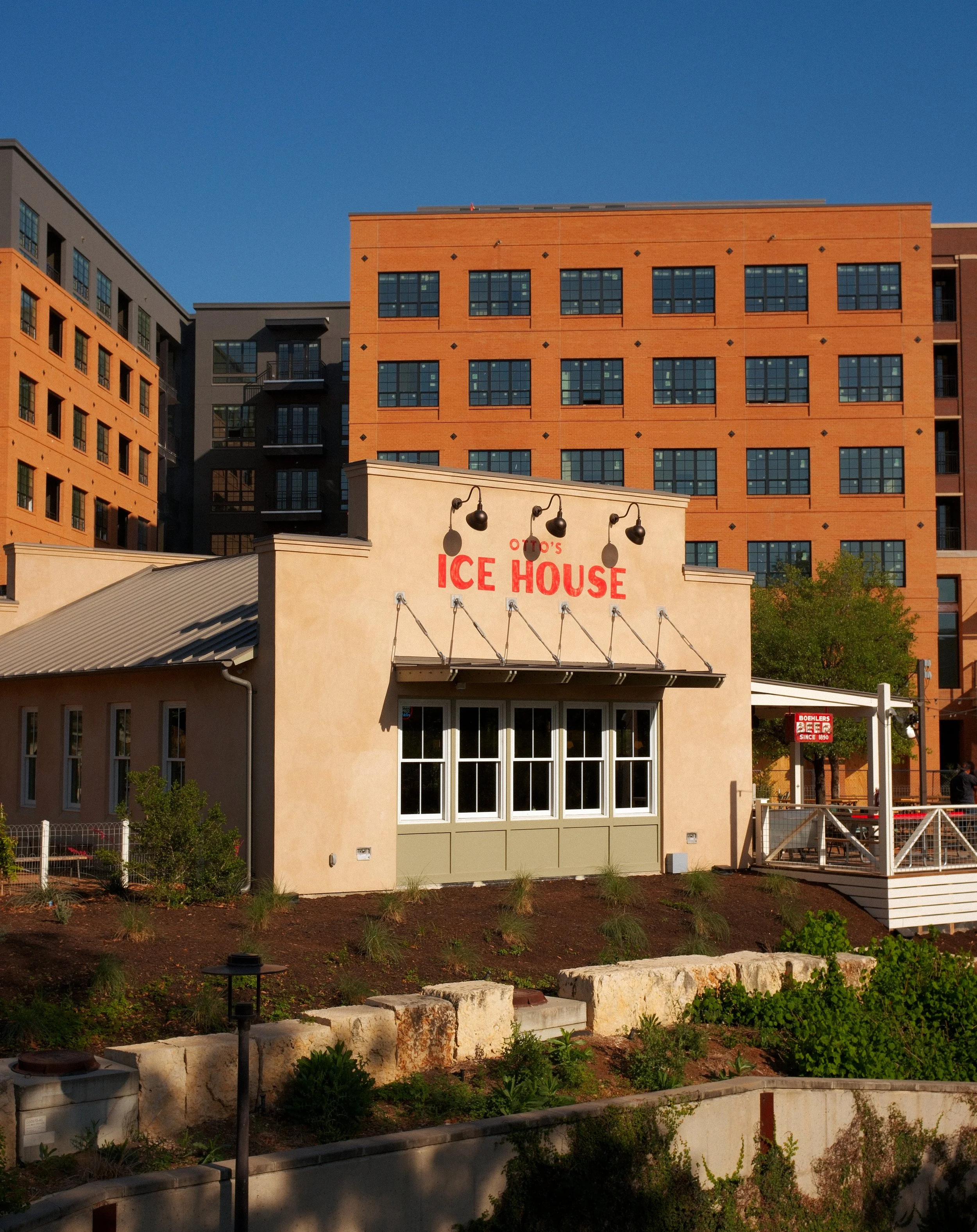 Small beige building with red sign that reads "OTO'S ICE HOUSE" in front of tall apartment buildings, with clear blue sky and some greenery.