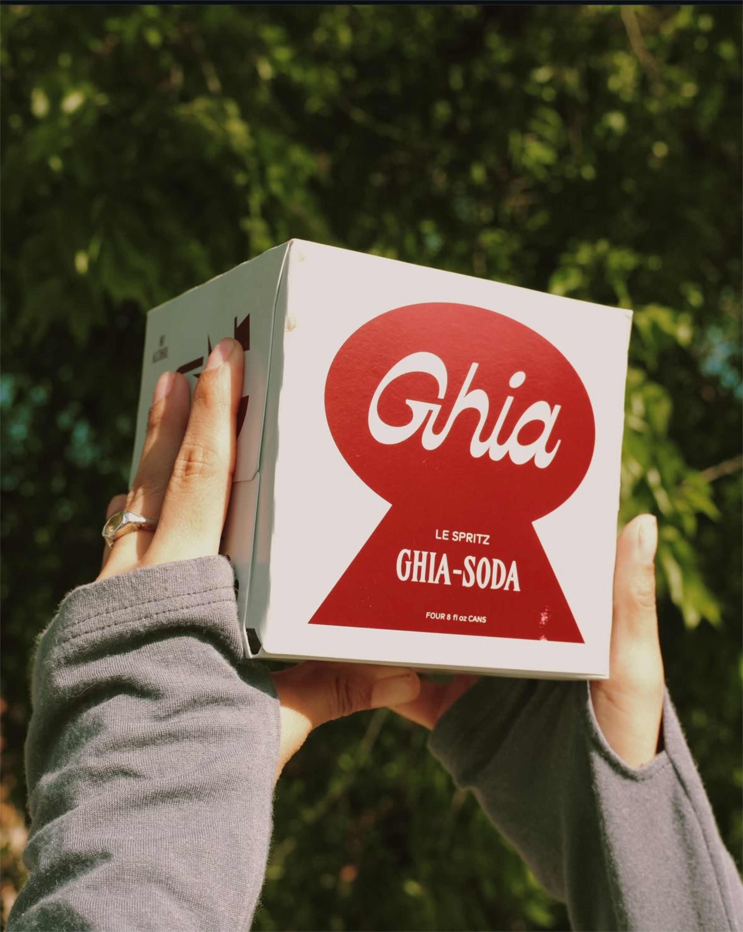 Person holding a box of Ghia soda outdoors with green trees in the background.