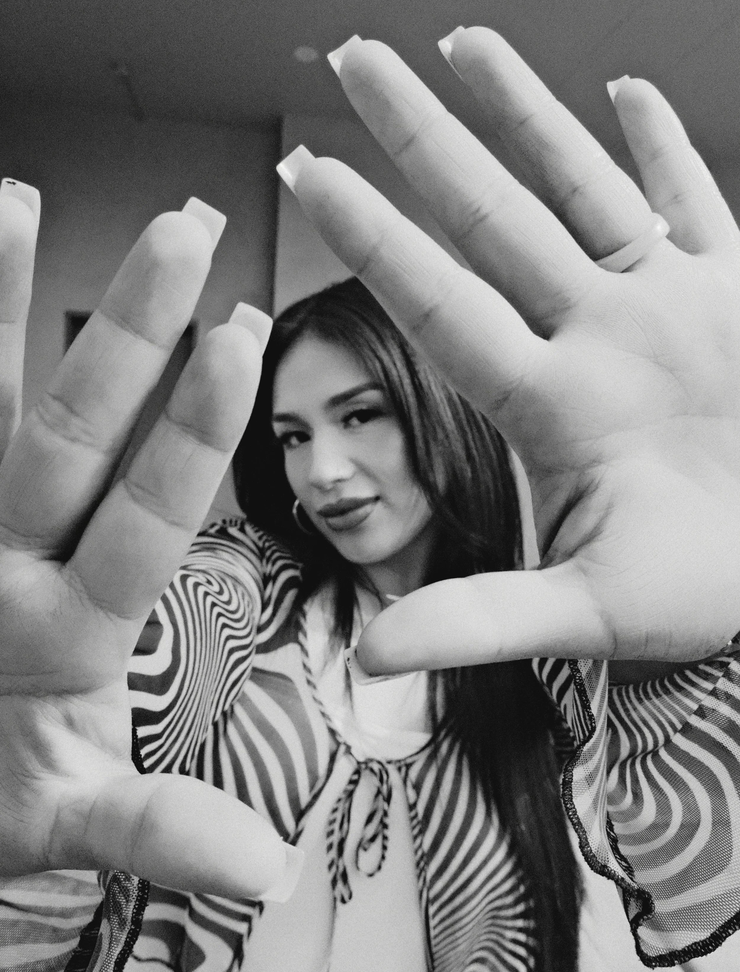 Black and white photo of a woman with dark hair, looking at camera, holding her hands up with fingers spread apart, in front of her face.