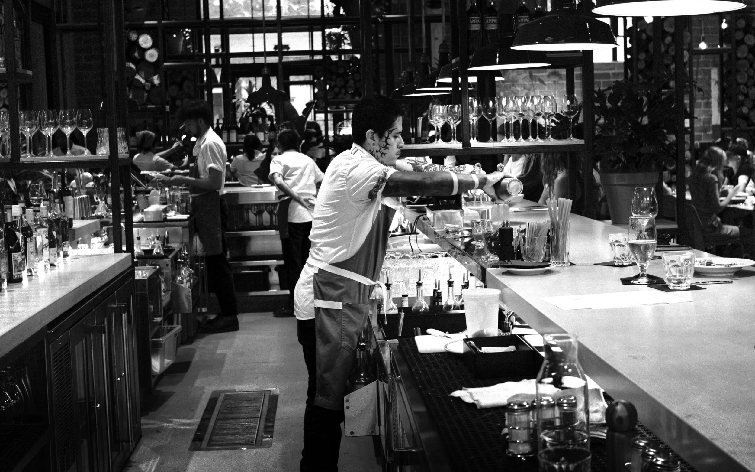 A busy restaurant bar with waitstaff preparing drinks and serving customers. The bar has various glassware and bottles, and customers are seated at the bar and tables.