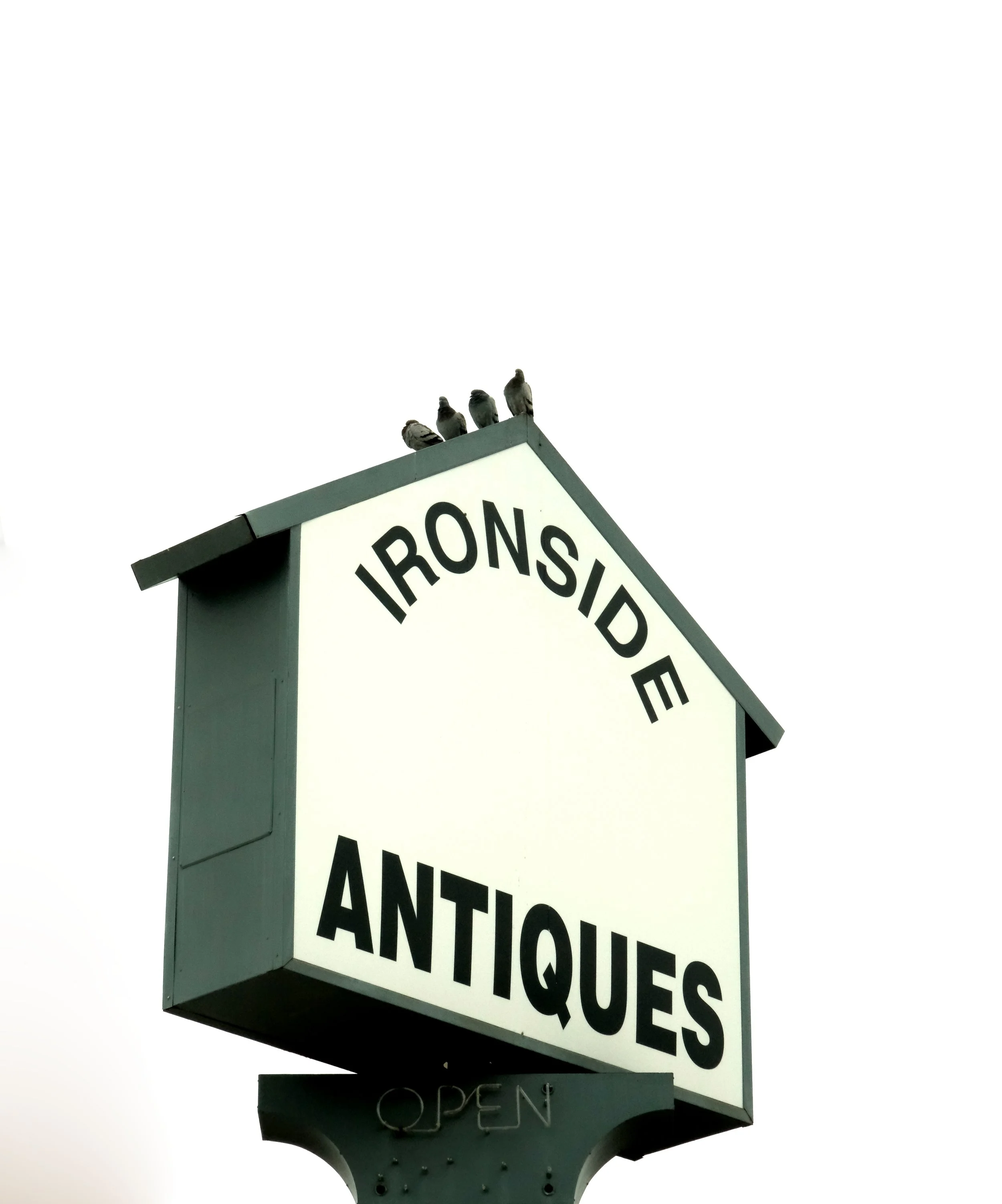 Sign for Ironside Antiques with the word 'OPEN' at the bottom, four pigeons perched on top of the sign, over a white sky.