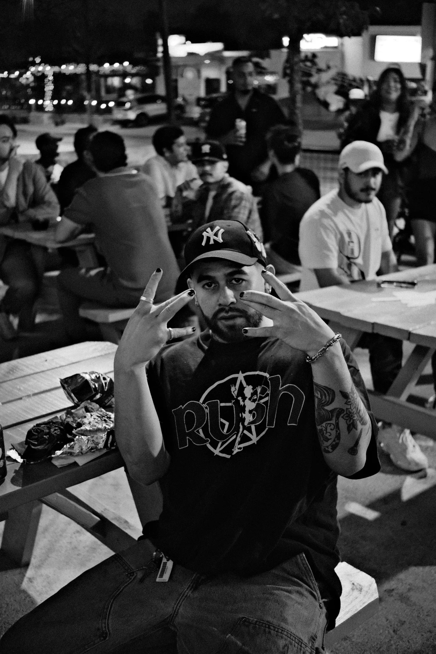 A man with tattoos and a beard wearing a 'Rush' t-shirt and a New York Yankees cap, sitting at a picnic table, making a gesture with his hands during an outdoor gathering at night.