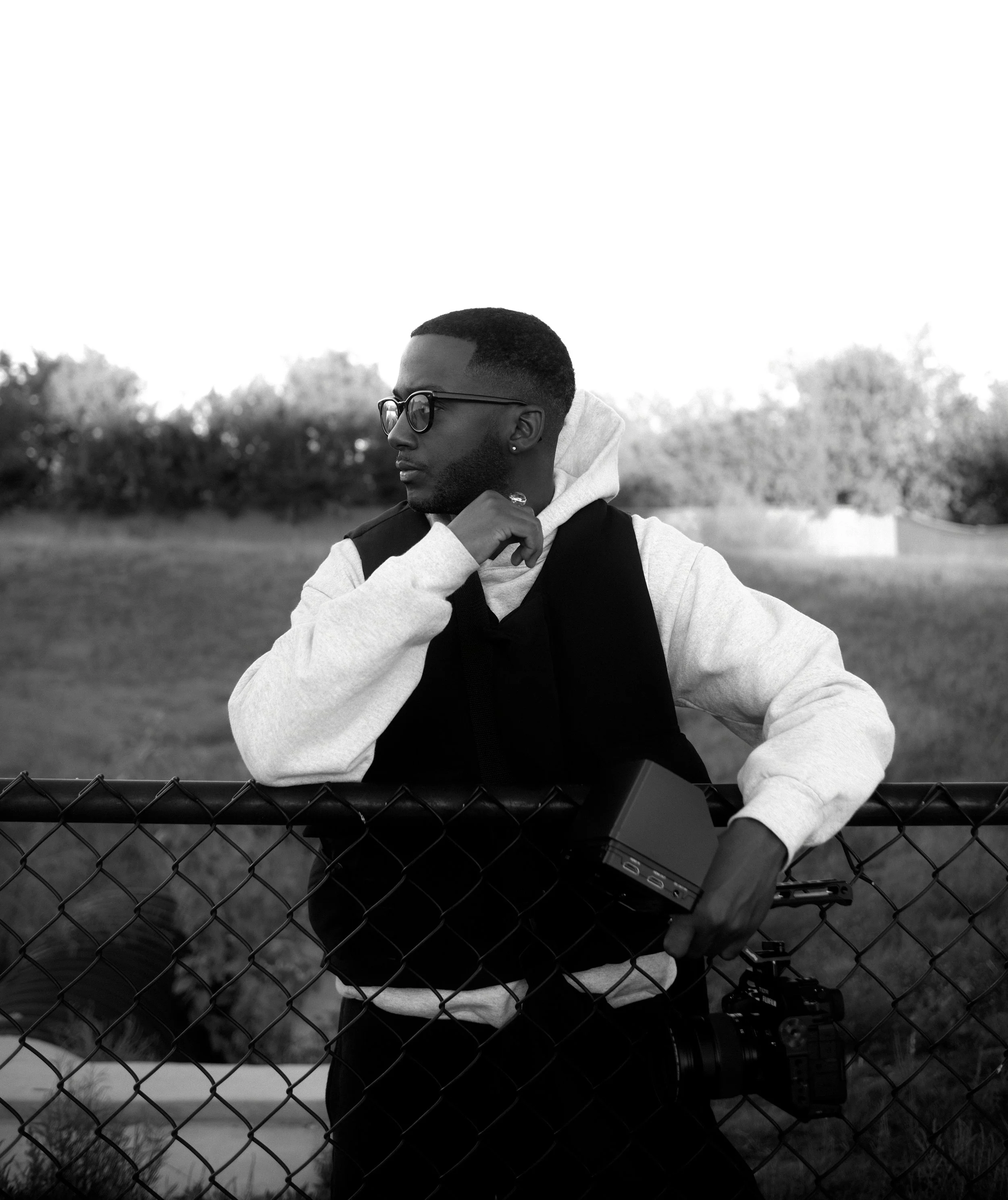 Young man with glasses leaning on a chain-link fence, wearing a hoodie and vest, holding a camera and a portable device in an outdoor park setting.
