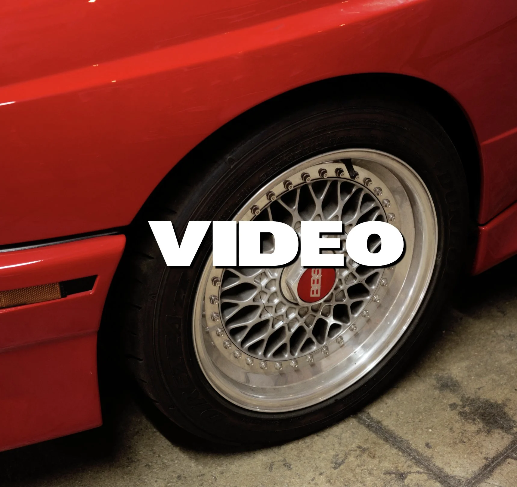 Close-up of a red sports car with a BBS alloy wheel and a black tire. The car is parked on a concrete floor.