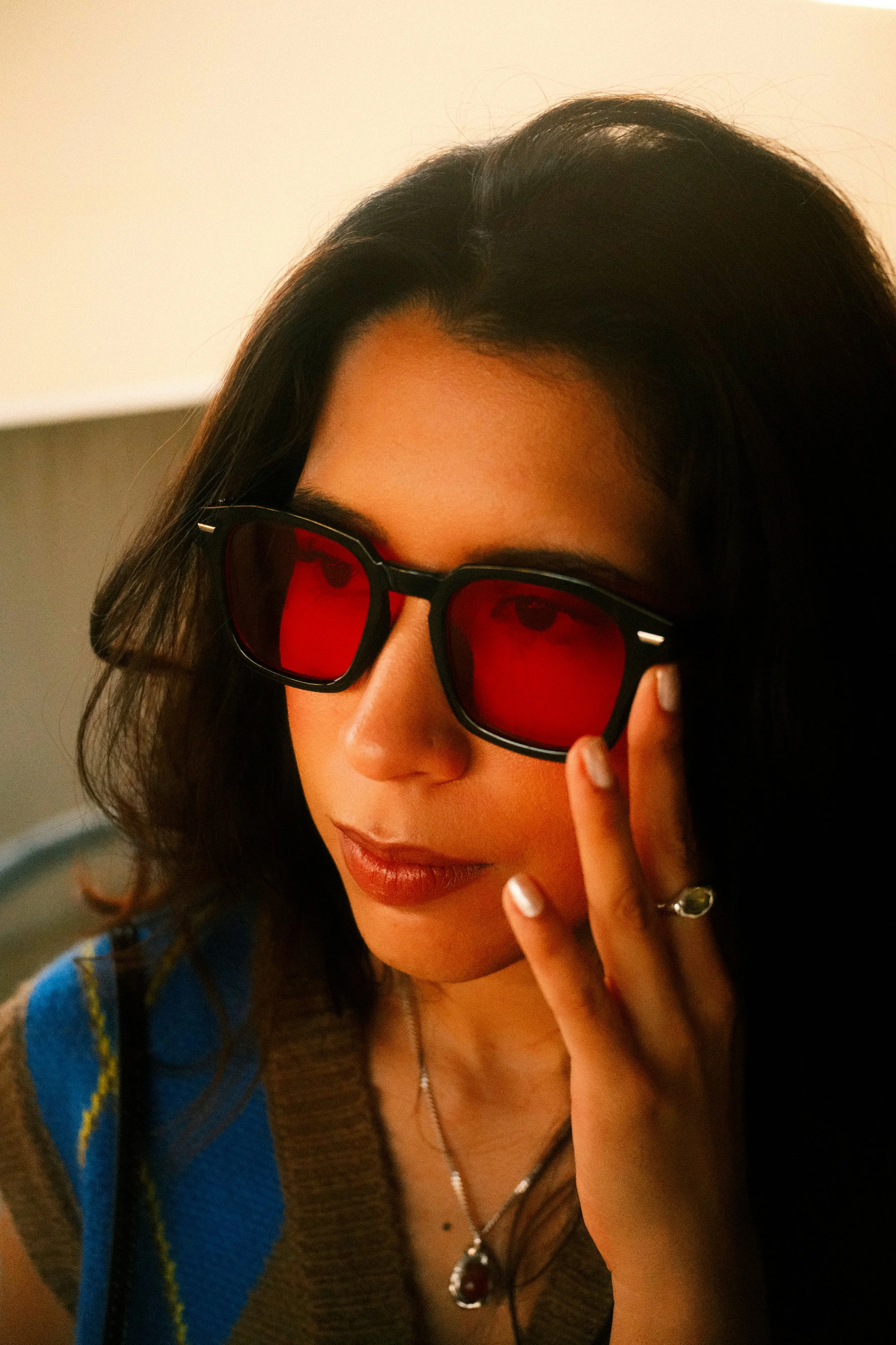 A woman with dark hair and light skin wearing sunglasses with red lenses, touching the frame of her glasses with her right hand, and wearing jewelry including a ring and necklace.