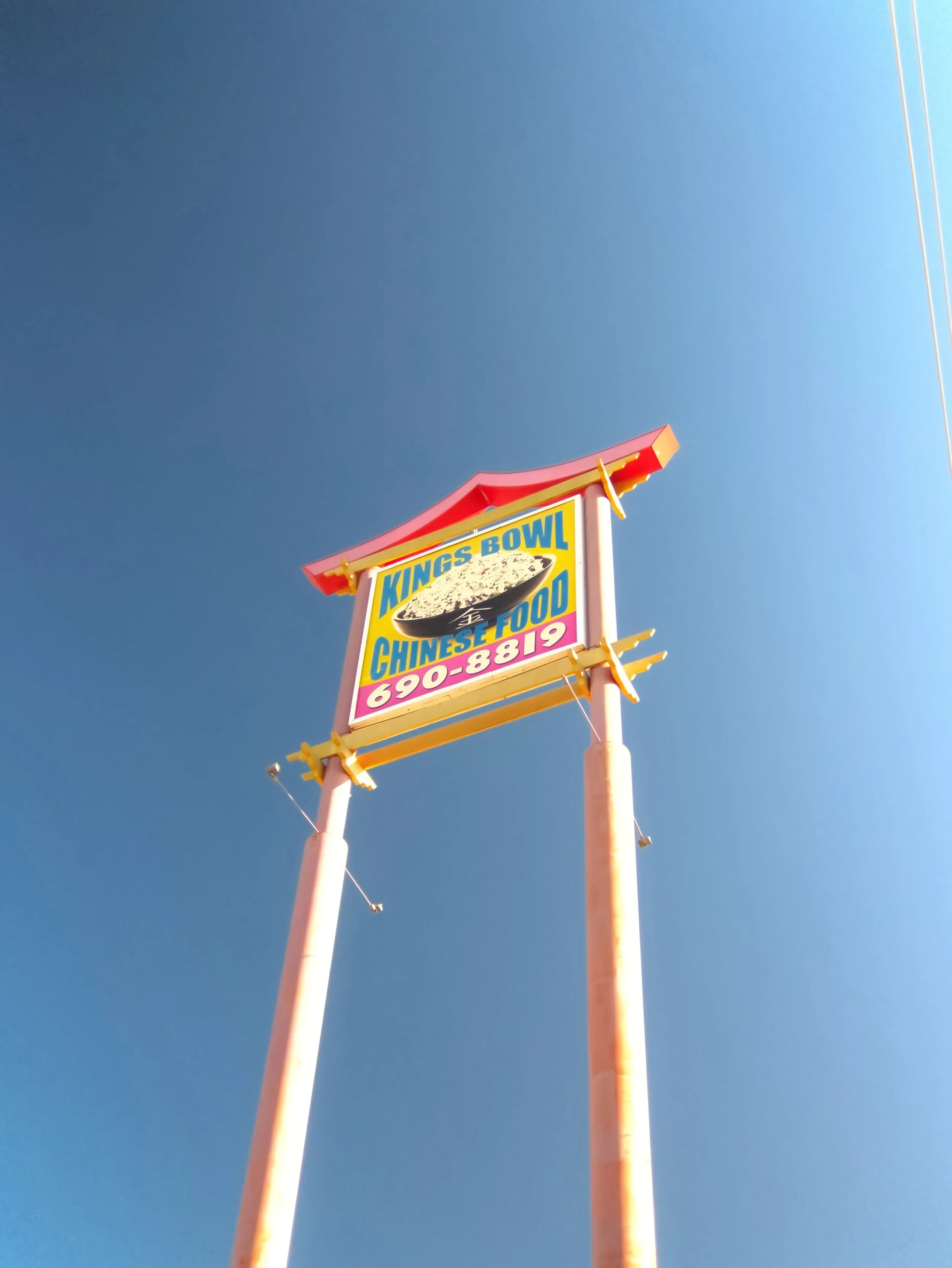 Sign for Kings Bowl Chinese Food with contact number, mounted on tall pink poles against a blue sky.
