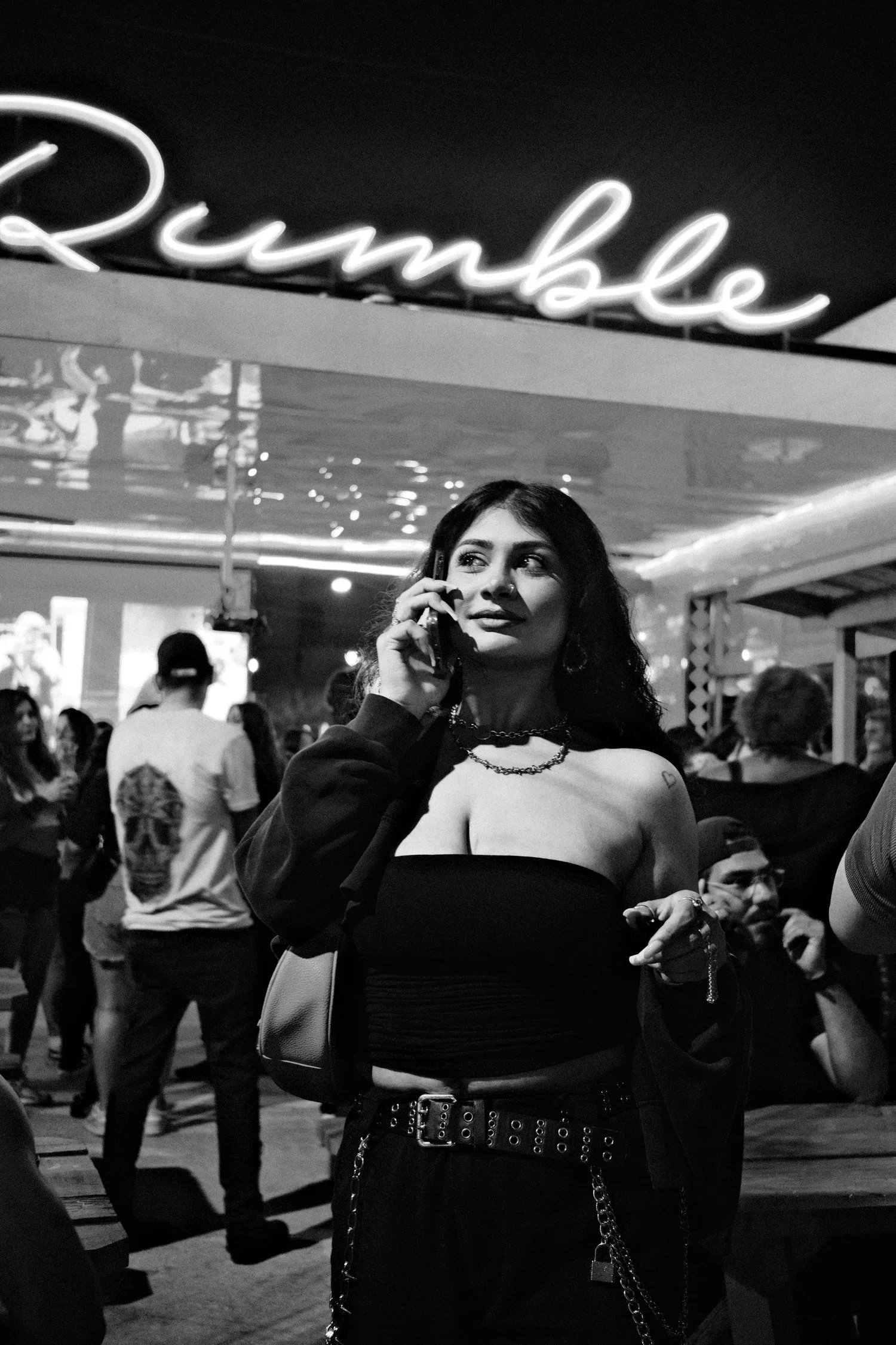 A young woman talking on a cellphone in a crowded indoor space, black and white photo, with a neon sign above her that reads 'Bounce'.