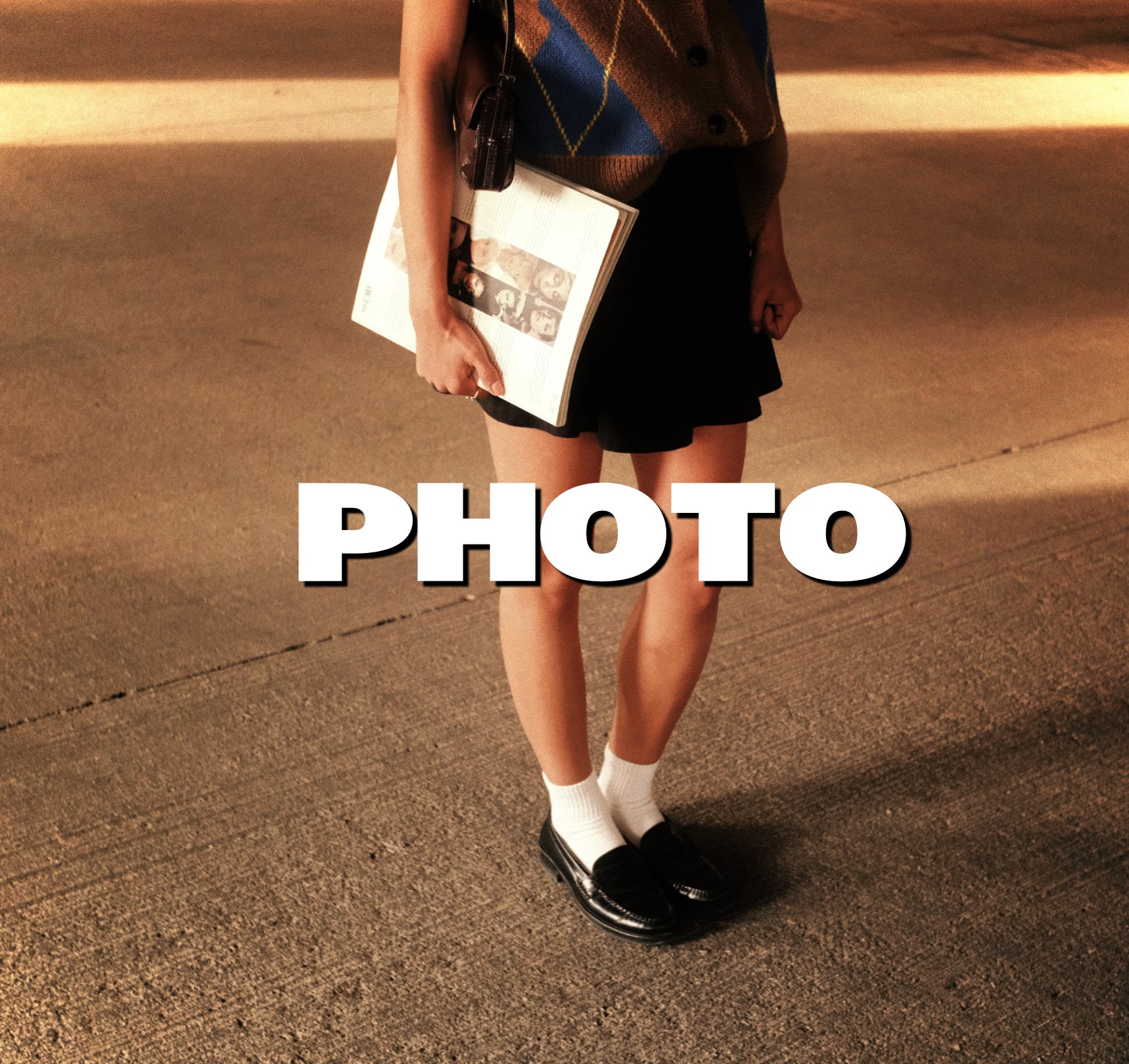 A person standing on a sidewalk holding a magazine or newspaper with a bag over their shoulder, wearing a plaid vest, a black skirt, white socks, and black loafers, with the word 'PHOTO' overlayed