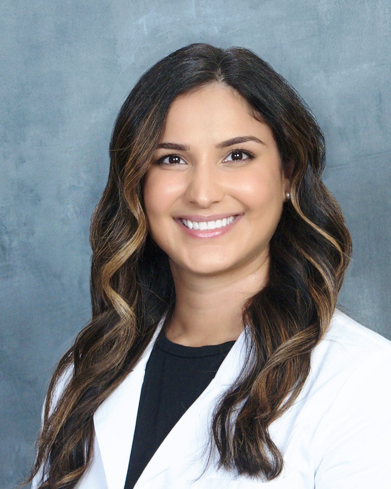 Portrait of a smiling woman with long wavy brown hair, wearing a white coat and black top, against a textured gray background.