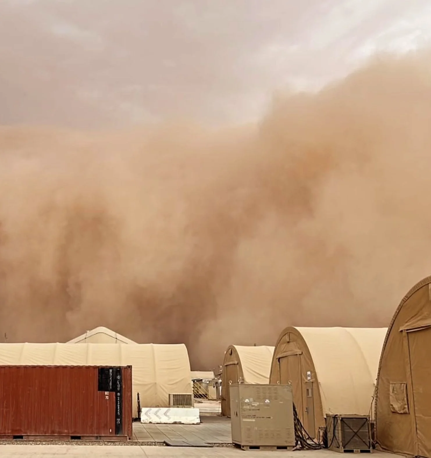 A row of beige military tents and a shipping container against a massive dust storm with dark, swirling clouds in the sky.