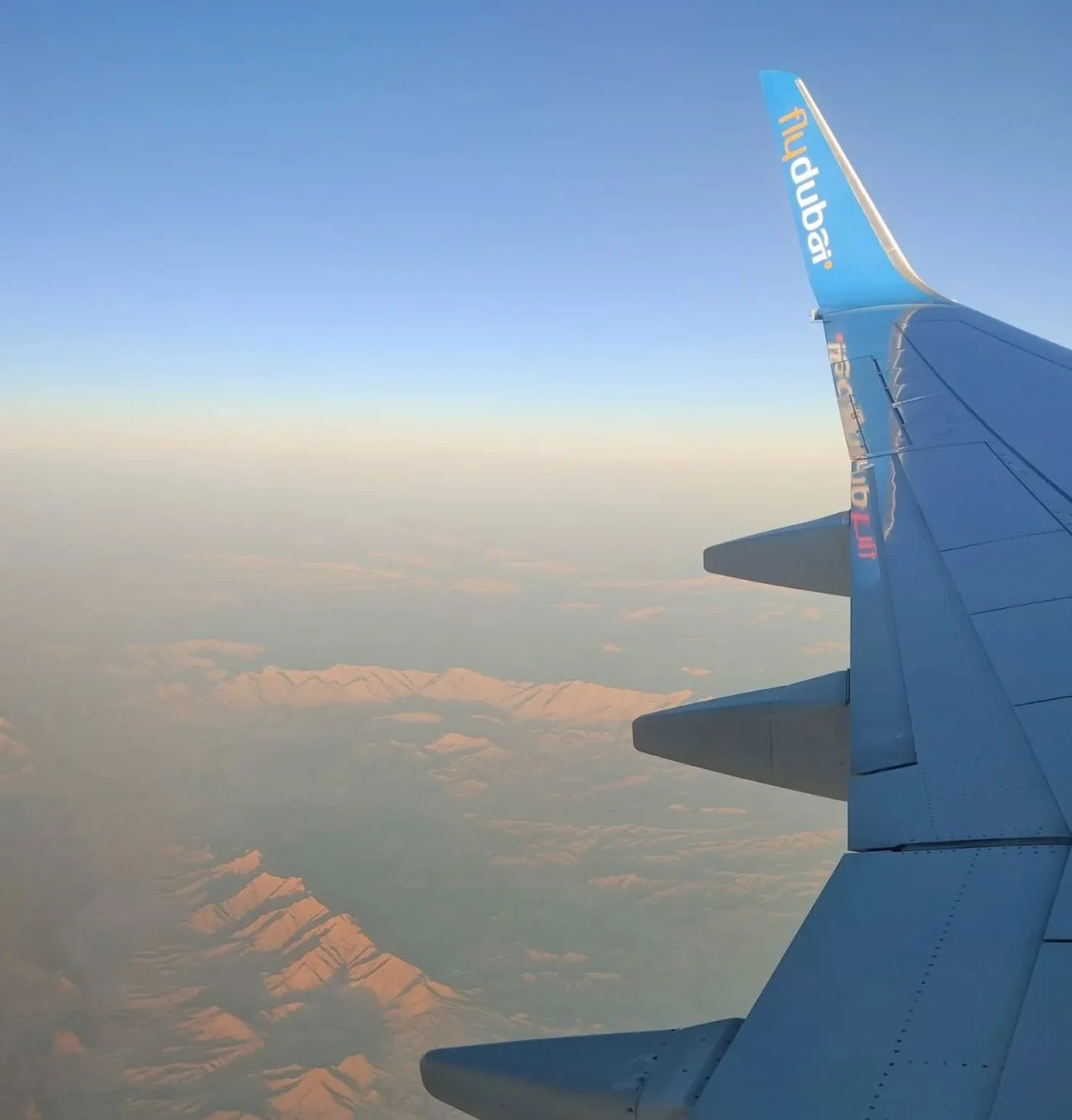 View from an airplane window showing a wing and mountain range below, with a gradient sky transitioning from blue to light near the horizon.