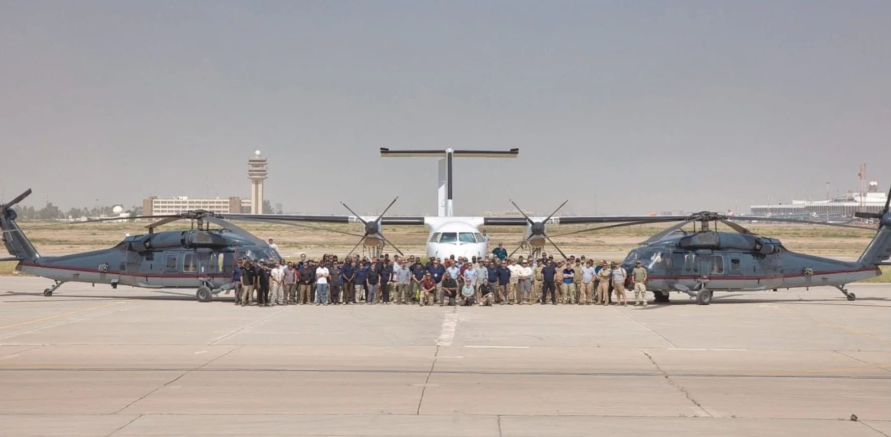 Group of people standing in front of two helicopters on an airport tarmac with a propeller aircraft in the background.