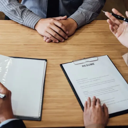 People in business suits sitting at a wooden table, with a pile of resumes and a clipboard with a resume on it in front of them, during a job interview.